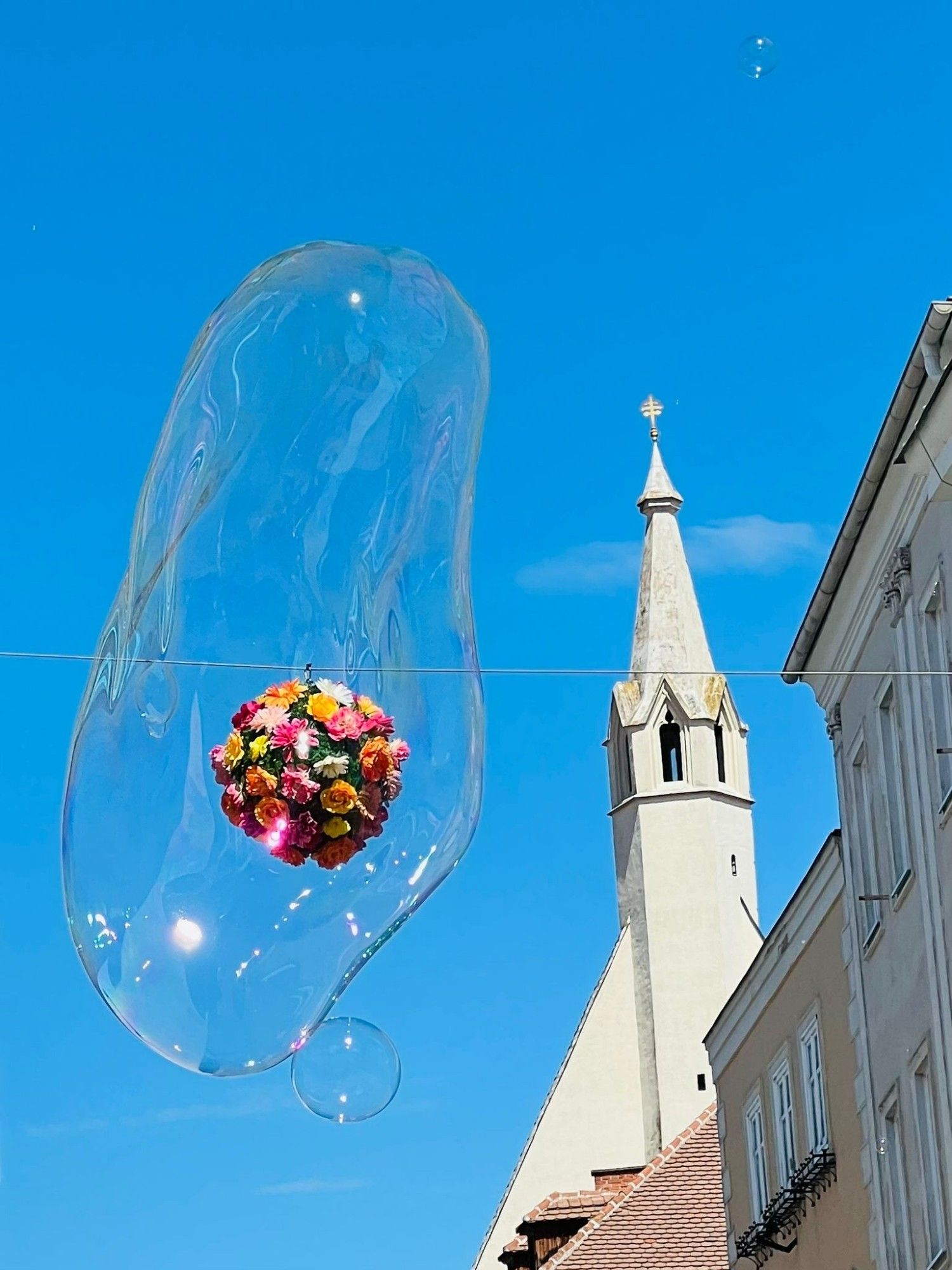 Hoch über der Fussgängerzone in Krems an der Donau hängt eine Blumenkugel als Deko, scheinbar umhüllt von einer Riesen-Seifenblase; rechts im Bild ein Steinkegel einer Kirchturmspitze