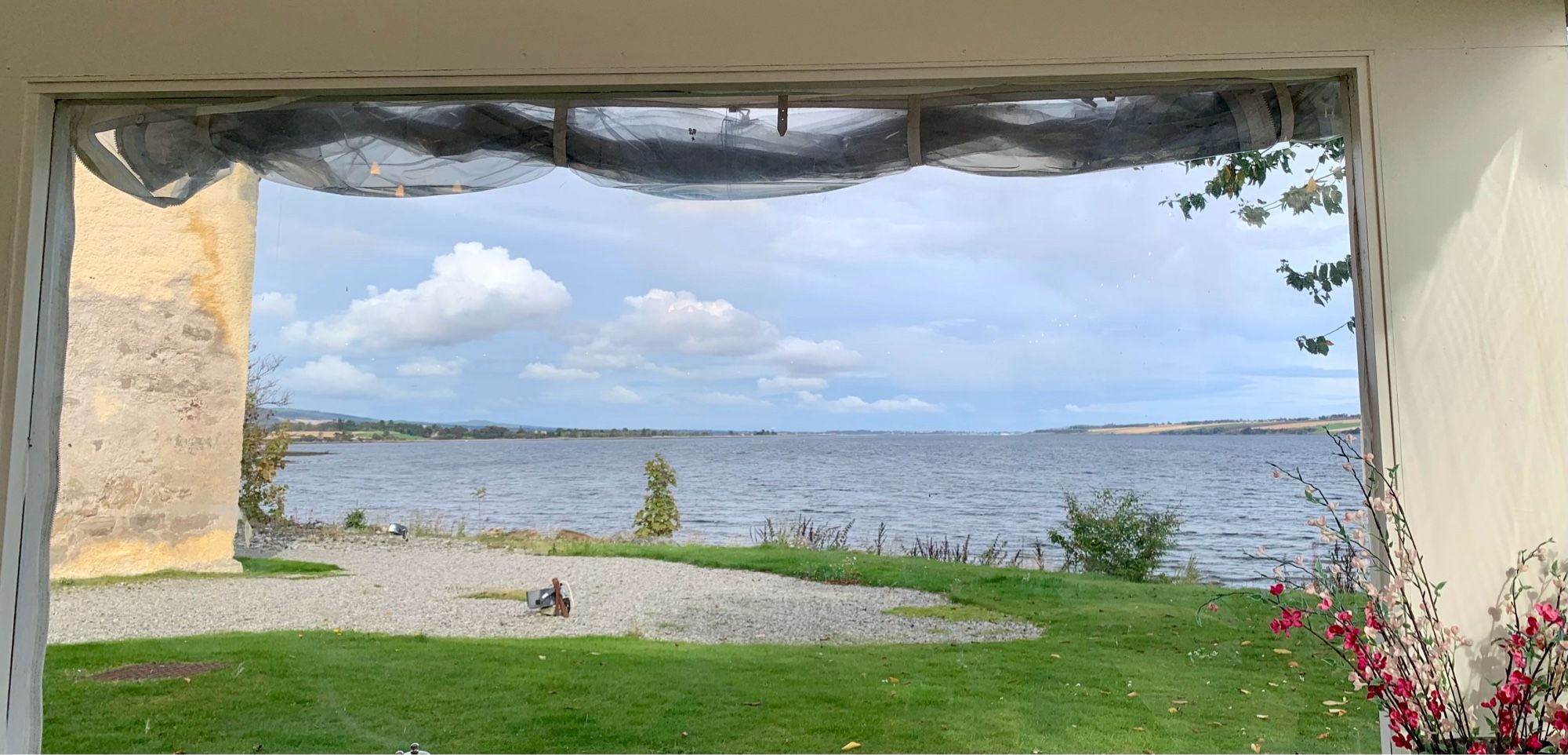 A view framed through a window opening in a marquee. Artificial flowers in the right corner.Some sort of netting pinned up above the top frame on the outside.Green grass, a gravel path, the corner of a building with deteriorated limestone covering. Water mid shot to the horizon. Land on both shores. White clouds in a pale blue sky.