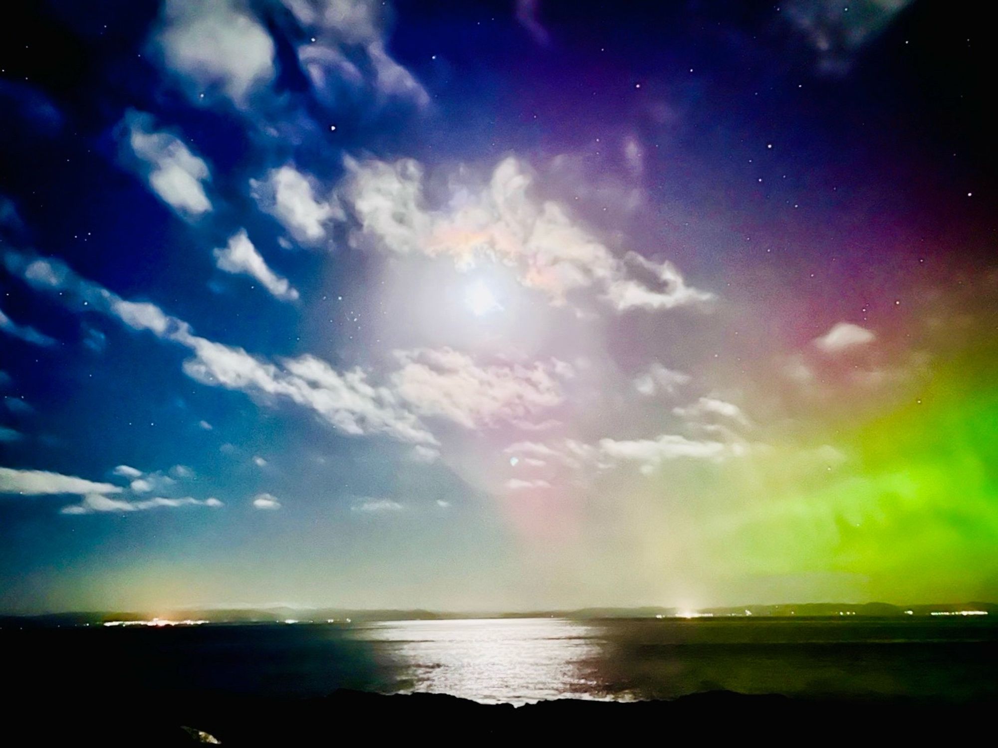 A cloudy sky with strong moonlight behind clouds. Aurora Borealis evident in purple and green casting right and centre. The moon is shining  over water giving a bright white reflection in the otherwise black surface, Lights on the horizon. Portmahomack Jan 2023. A photo by D Ross