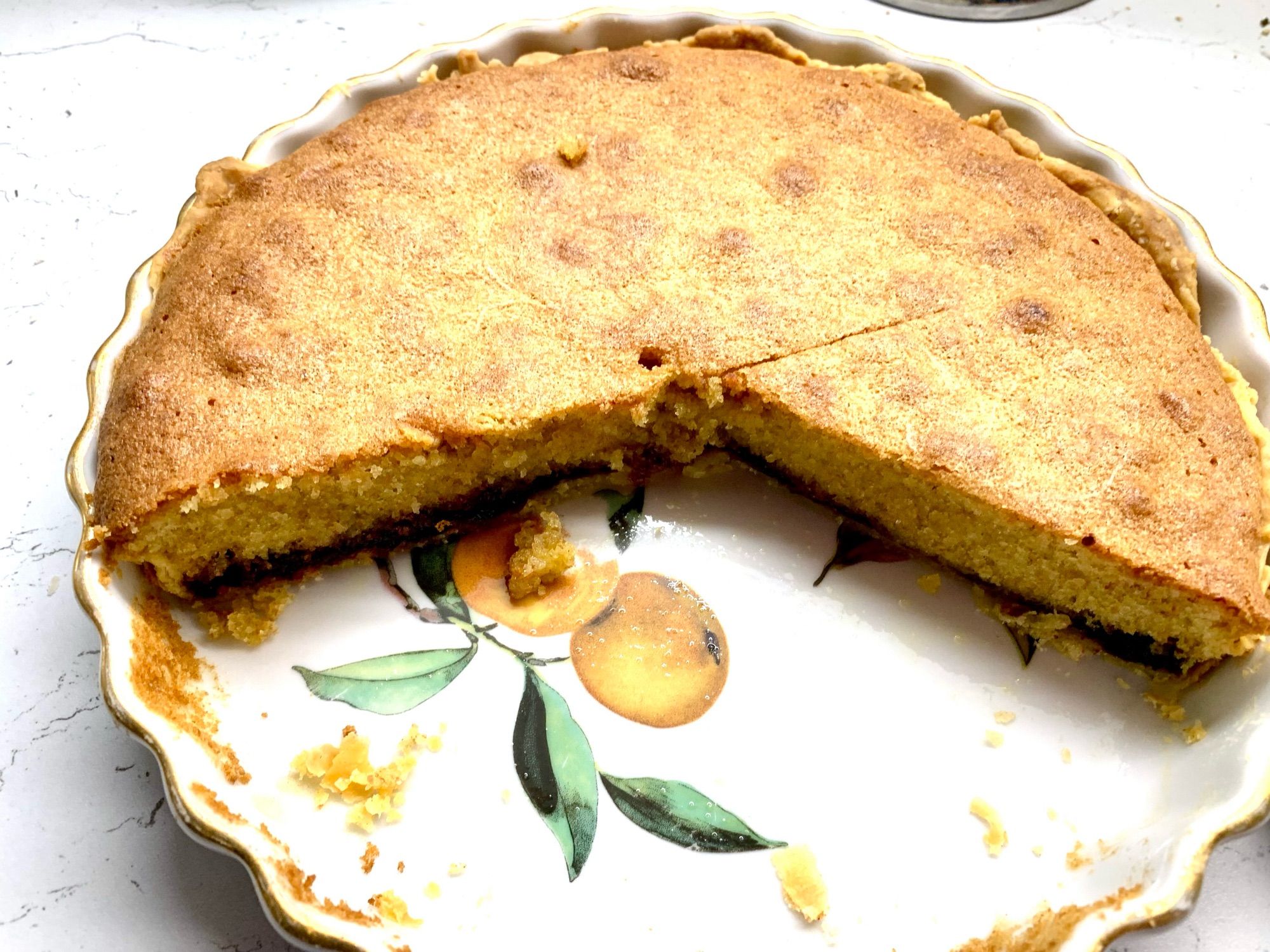 Fluted flan dish with a design oranges on the base. 3/4 of a tart inside with jam bottom and frangipane top. Pastry visible on the rim.