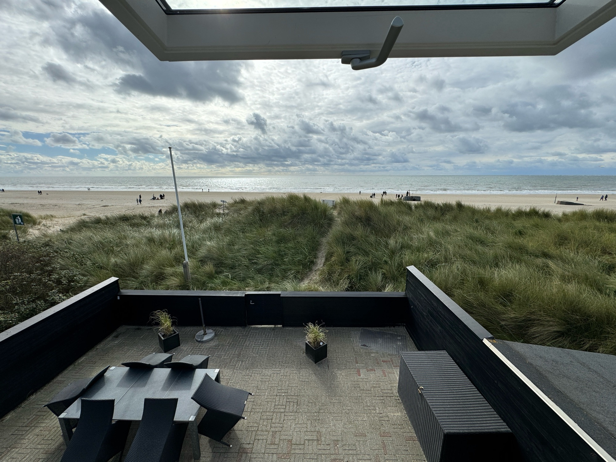 Blick vom Haus auf das Meer. davor ist eine bewachsene Sanddüne. Wiederum davor sieht man die Terrasse vom Haus. Darum herum ist ein schwarzer Zaun. man sieht links einen Esstisch mit sechs Stühlen, rechts eine Outdoor Kiste. Was man nicht mehr sieht, ist der Grill