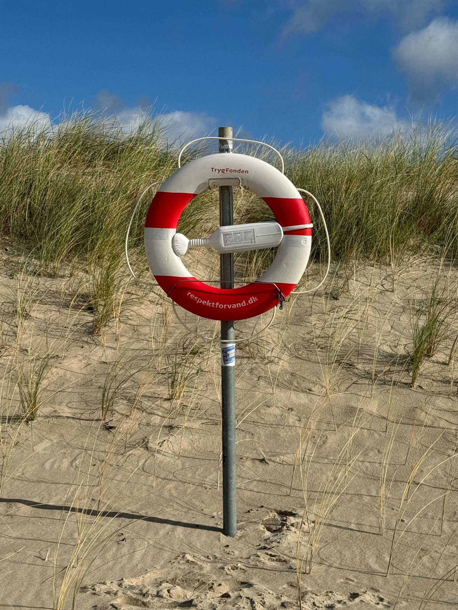 Ein Rettungsring hängt an einer Stange. Darunter ist Sand. Dahinter sieht man die Düne mit grünem Bewuchs. Es scheint wunderbar die Sonne