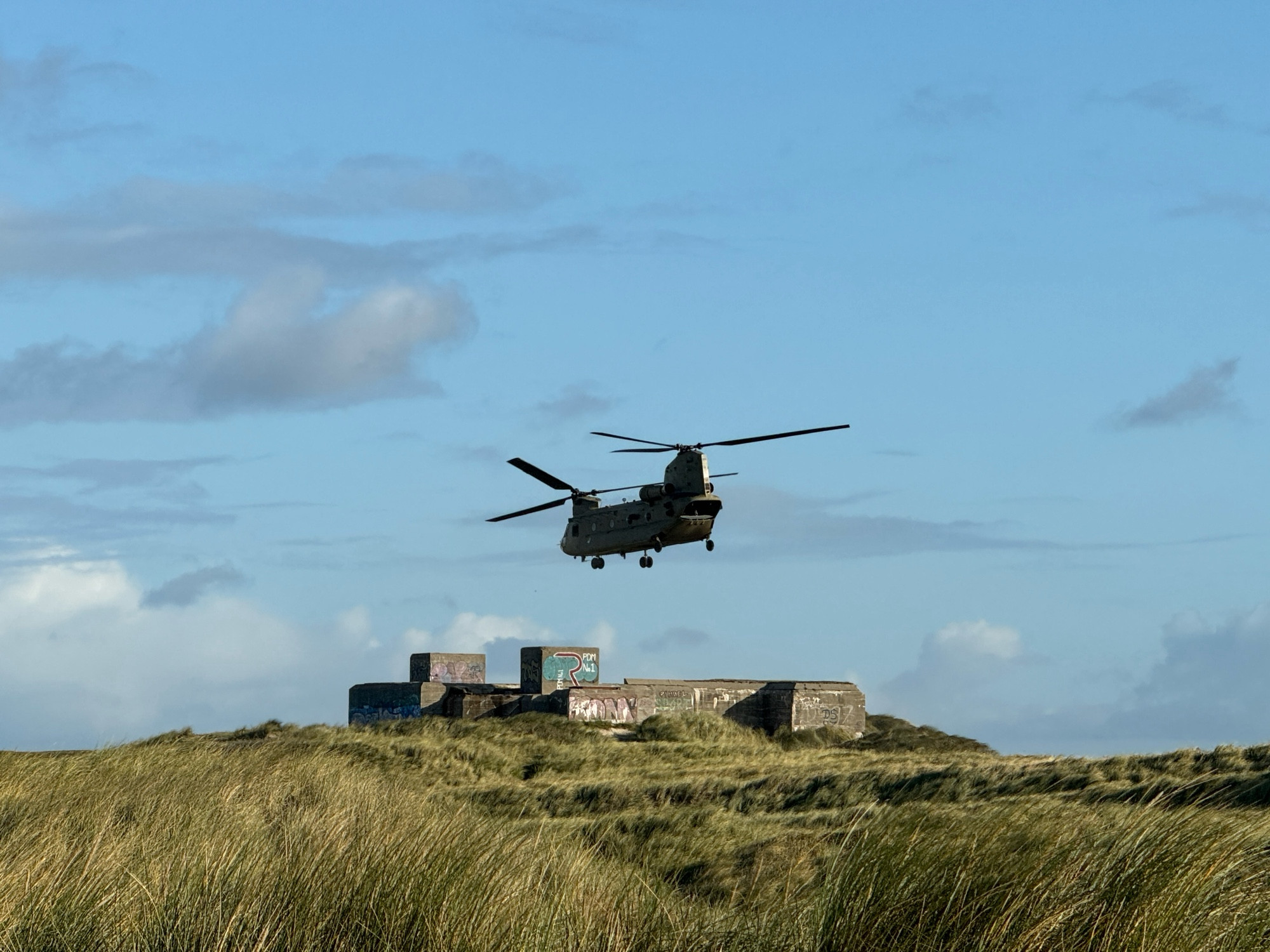 Deutscher Bunker vom Atlantikwall aus dem Zweiten Weltkrieg. Darüber ein Helikopter der dänischen Armee im Vordergrund Dünengras. Der Himmel ist blau mit Wolken.