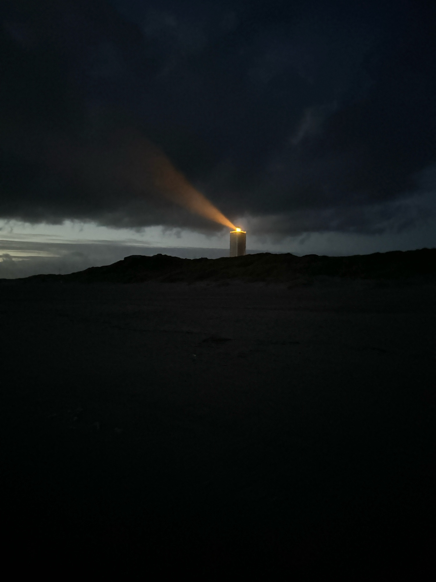 Bild ist fast schwarz-weiß, weil im Dunkeln aufgenommen. Man sieht den Leuchtturm im Hintergrund. Der Leuchtturm steht in einem Bereich, in dem ein hell grauer Streifen quer durch das Bild geht. Es handelt es sich um den Horizont. Darüber sind dunkelgraue Wolken, darunter und links mittelgrau das Meer,  vorne ist der Strand und die Dünen. Alles in schwarz. Man kann noch ein bisschen Schemen erahnen