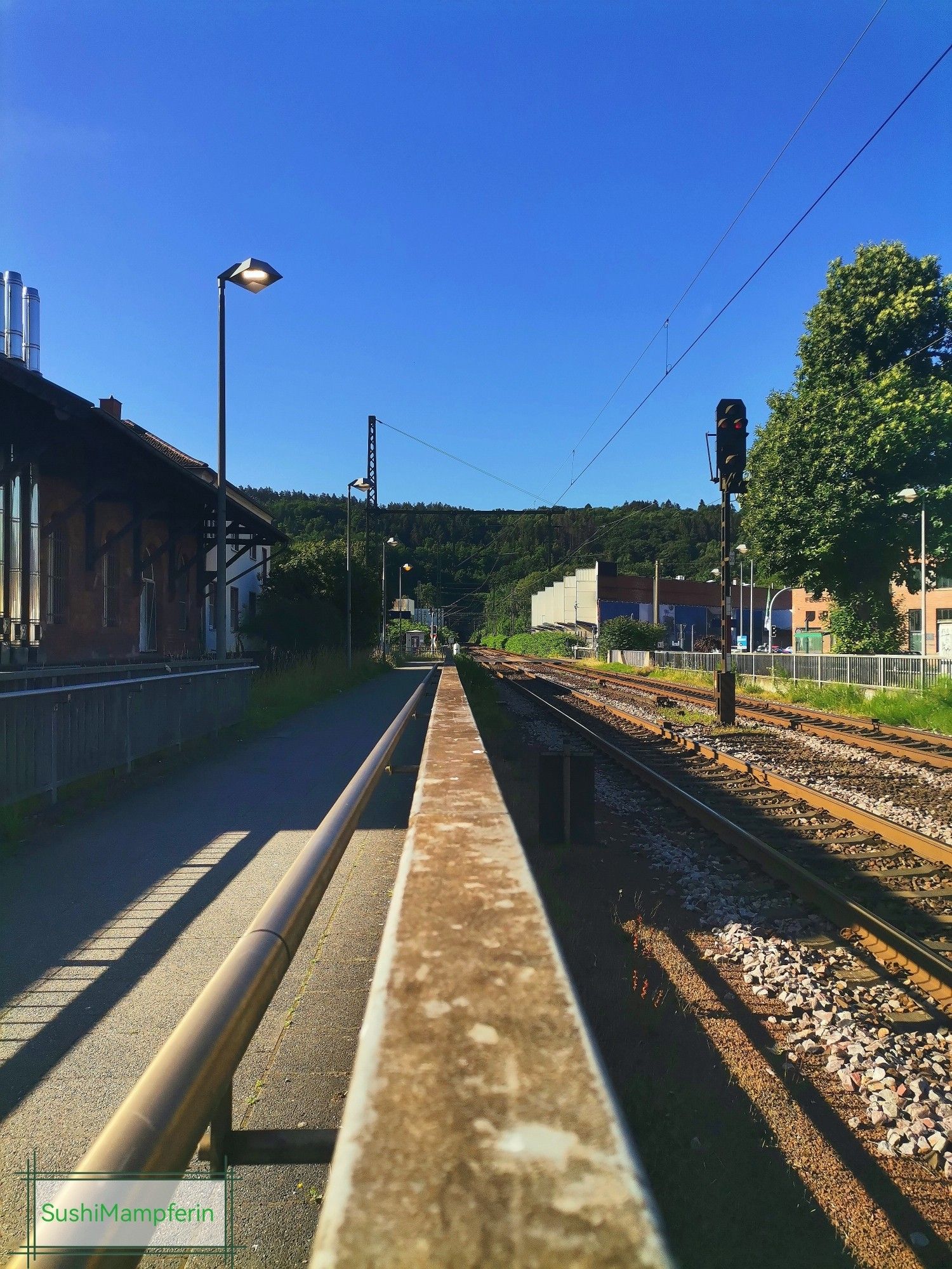 A picture of a trainstation. To the left side you can see a little building, on the right side the rails. The sky is bright blue, sunny day.