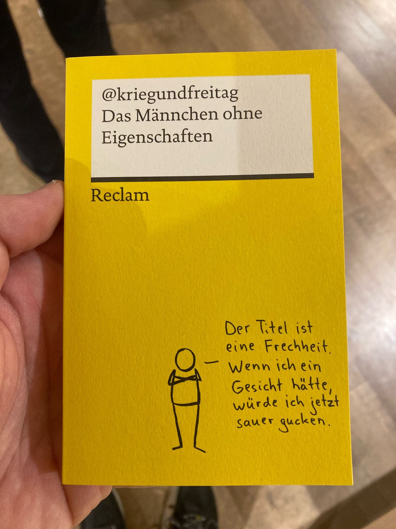 Im Bücherladen stehend, Großaufnahme meiner Hand, das Buch „Das Männchen ohne Eigenschaften“ von @Kriegundfreitag haltend.