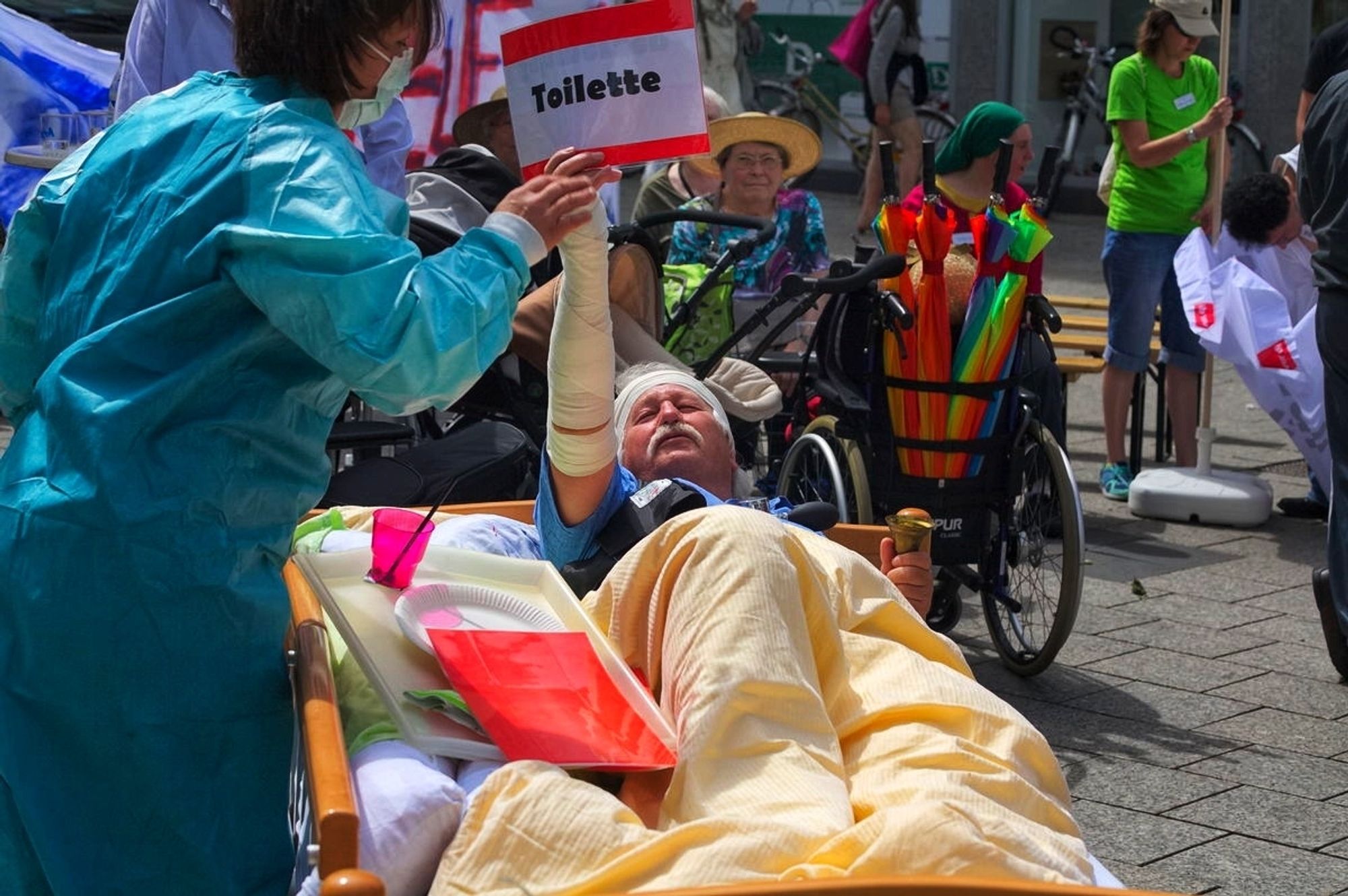 Das Bild entstand bei der Demo "Tatort Pflegenotstand Ulm"
Das Bild zeigt einen Mann mit Kopf -und Armverband in einem Pflegebett. Das Bett steht in der Fußgängerzone. Er hält verzweifelt ein Schild in die Höhe, auf welchem "Toilette" steht. Eine Schwester im OP-Kittel jetzt durch die Szene und vertröstet den Patienten.Im Hintergrund sieht man weitere Demo - Teilnehmer.