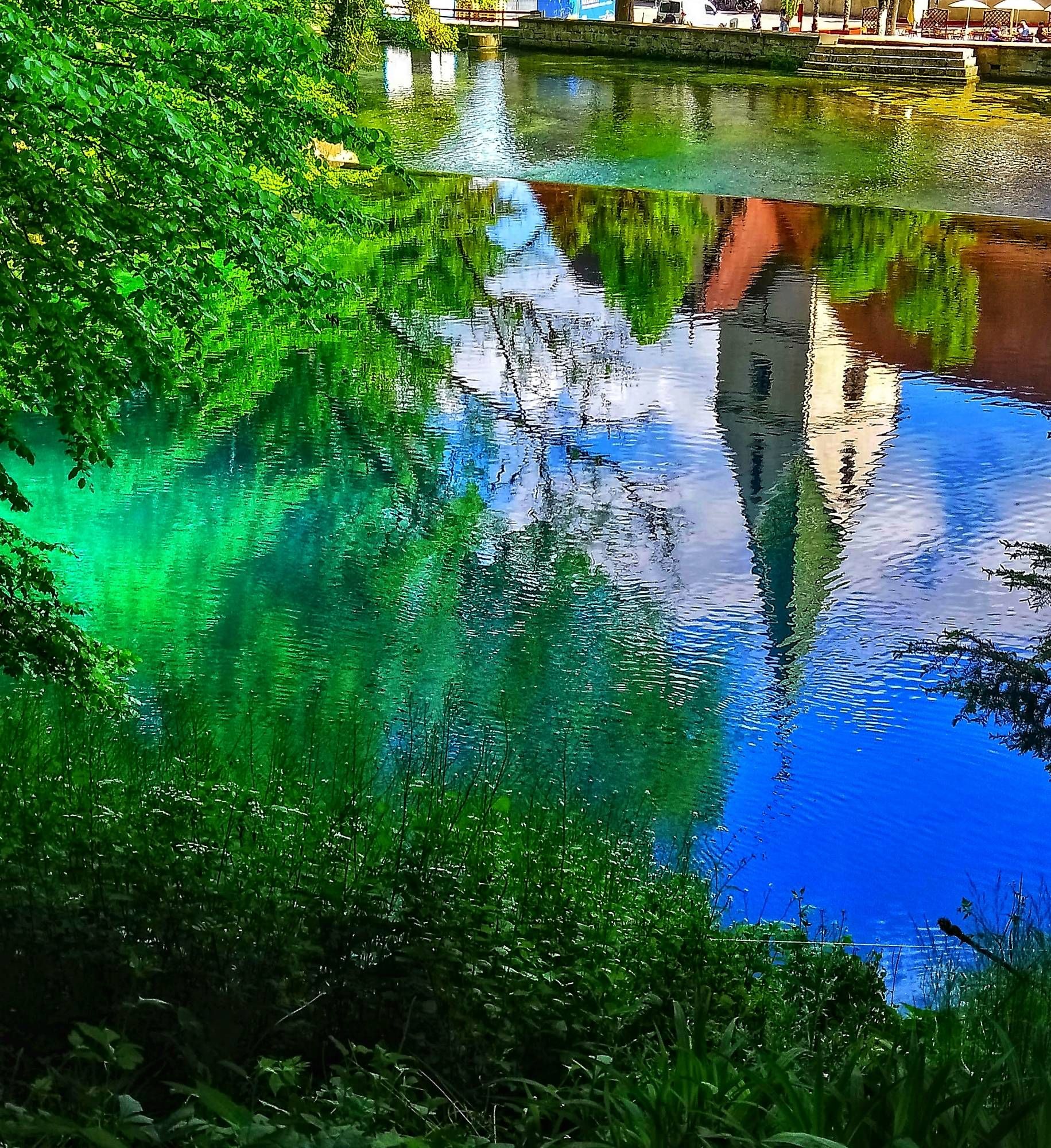 Das strahlend blaue Wasser des Blautopf (Quelle des Flüsschen Blau, in Blaubeuren bei Ulm). Im Wasser spiegelt sich ein Kirchturm mit spitzem grünen Dach. Am linken und unteren Bildrand sieht man Bäume und Sträucher, die sich ebenfalls im Wasser spiegeln.