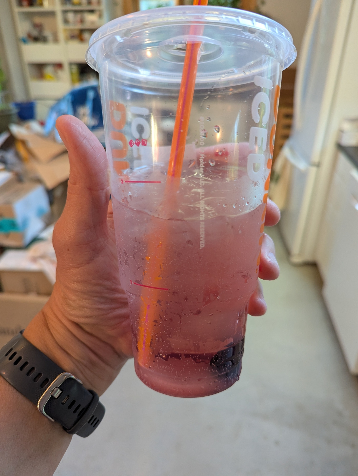 Hand holding up a large Dunkin Donuts beverage cup containing an iced, mostly clear liquid with maraschino cherries visible on the bottom imparting a pink tinge