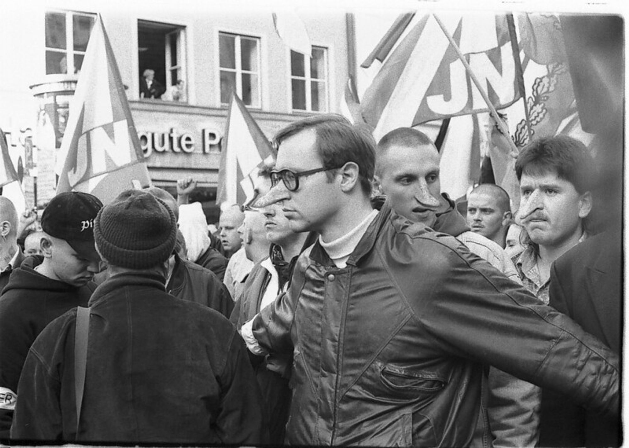 Die SW-Aufnahme zeigt mehrere männliche Teilnehmer einer Demo der Nazi-Organisation "Junge Nationaldemokraten". Im Hintergrund eine ältere Hausfassade, wo an einem geöffneten Fenster 2 Personen stehen. Die Nazis halten mehrere Fahnen mit "JN"-Schriftzug. Vorne rechts steht ein Nazi mit dunklem dünnen Blouson, Brille mit dunkler Fassung und dunklen Haaren. Er hat die Arme ausgebreitet, als wolle er die Menge hinter ihm zurückhalten. Ihm und zwei Nazis direkt hinter ihm, davon einer mit rasiertem Kopf, der andere mit Vokuhila und Schnäuzer, wurden mittels Photoshop die Nasen langgezogen.