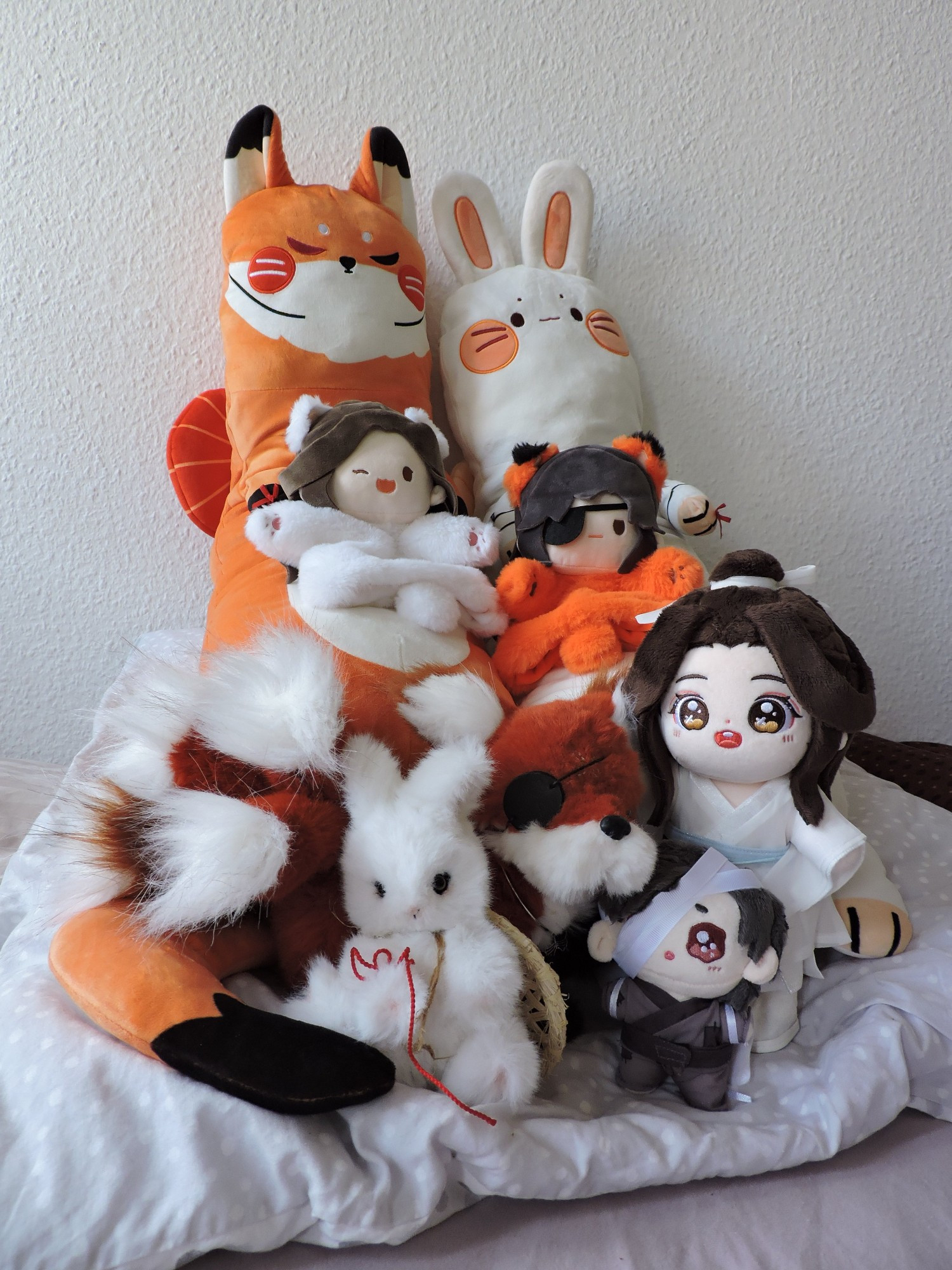 Photo of eight plushies representing Hua Cheng and Xie Lian from the novel Heaven Official's Blessing in different shapes, organised together atop a big square pillow.