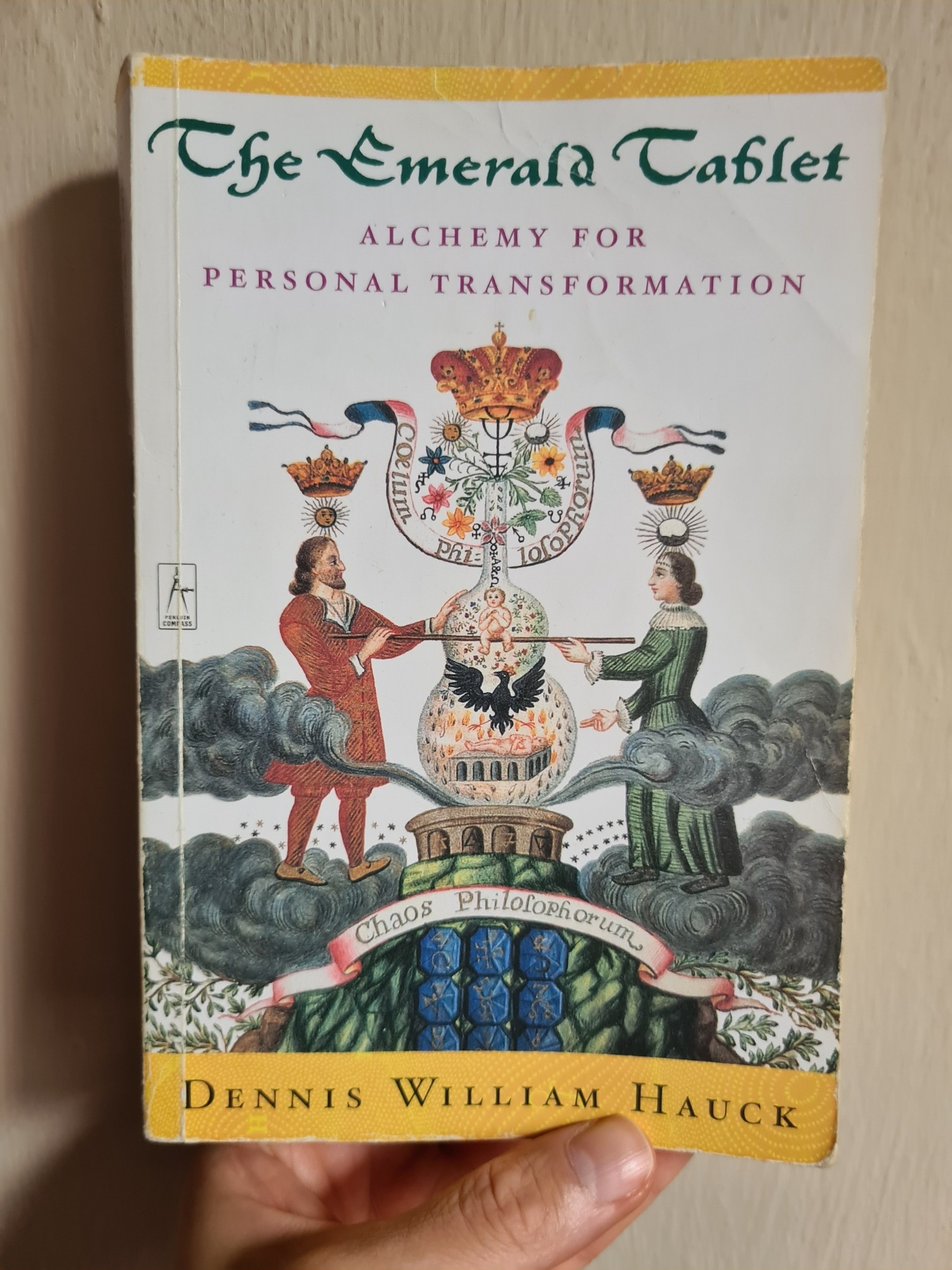 Thea's hand holding up a copy of Dennis William Hauck's book, 'The Emerald Tablet: Alchemy for Personal Transformation'.