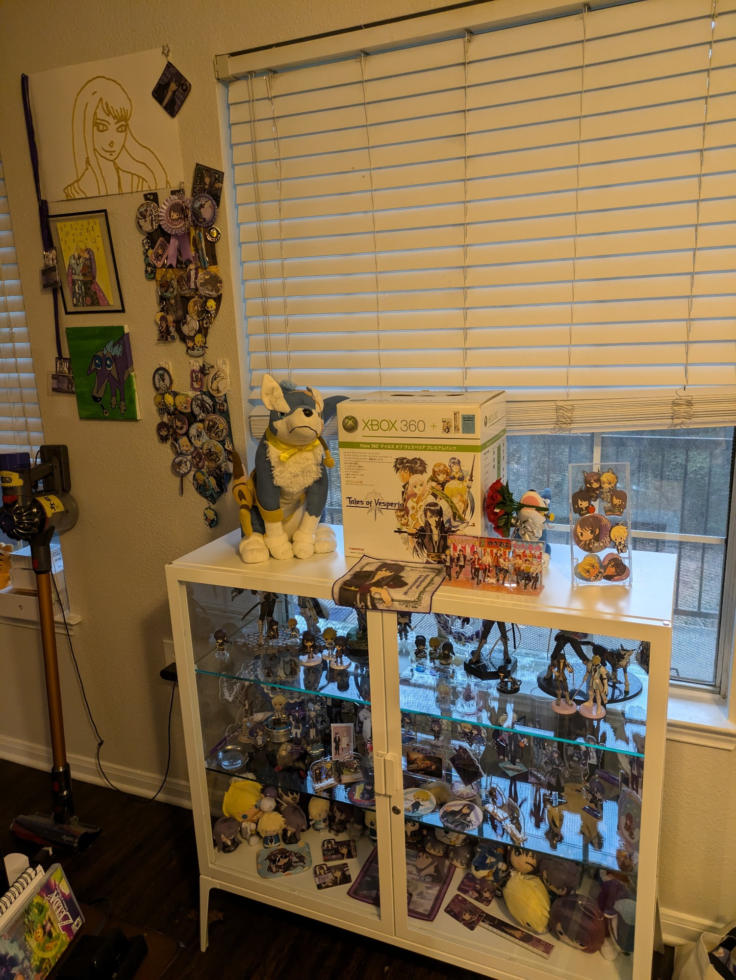 a display case and wall containing official and fan merchandise/art of the characters yuri lowell, flynn scifo, and repede from the game tales of vesperia. notable items are a large plushie of repede, the official tales of vesperia xbox 360, and macaroni art of yuri lowell.