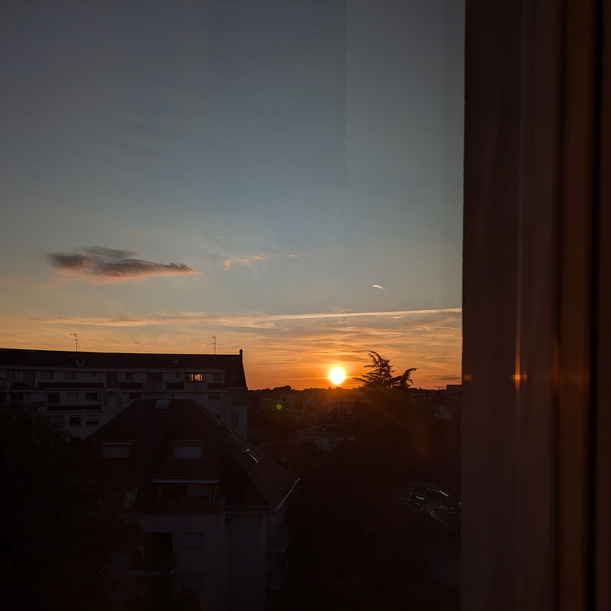 Photo d'un coucher de soleil flamboyant prise de derrière une fenêtre. Le soleil, orange, est bientôt derrière la ligne d'horizon, au 1er plan on distingue les silhouettes sombres d'un immeuble et d'arbres. Le ciel est bleu-gris, de rares nuages parsèment le ciel.