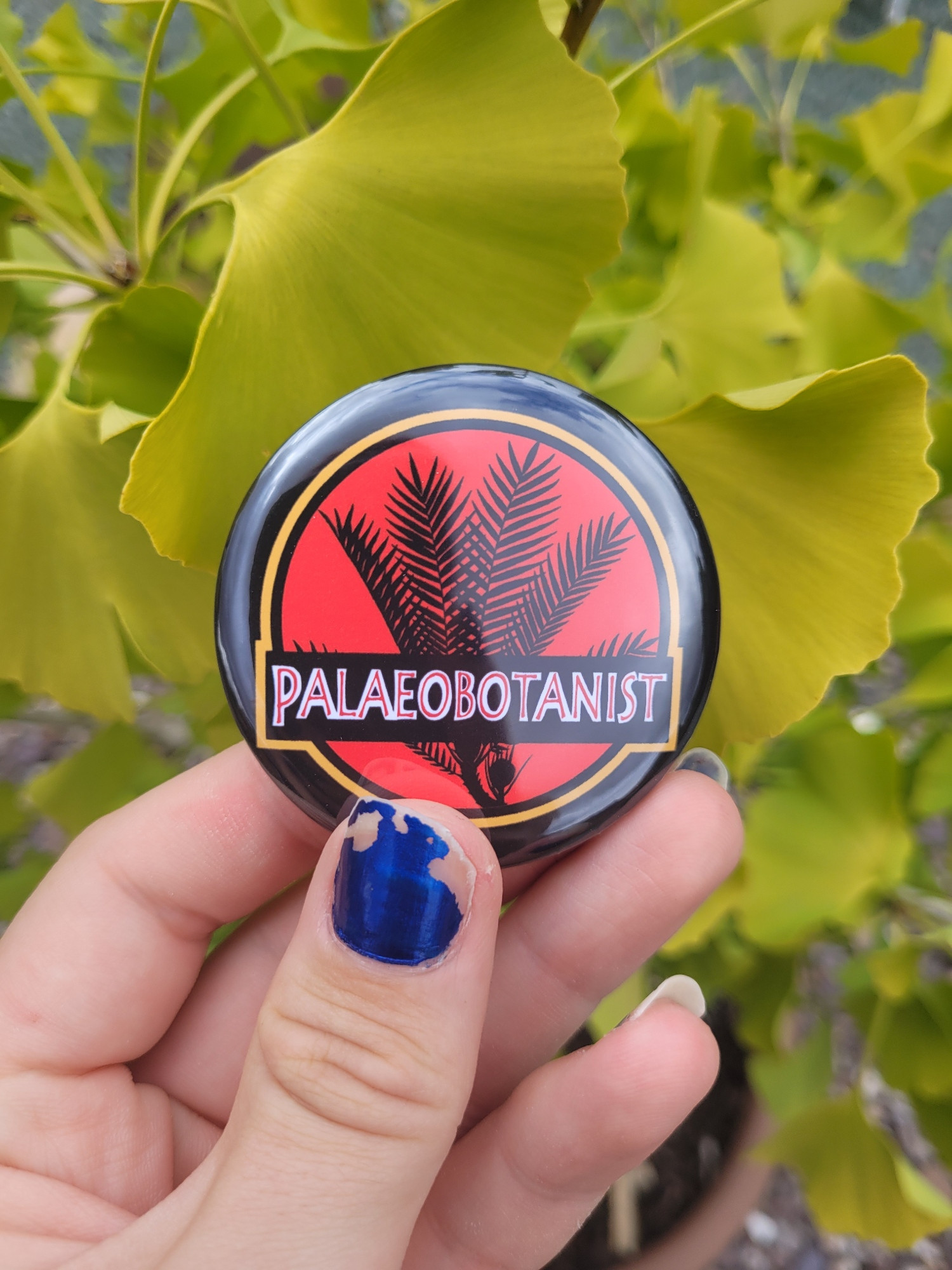 The Jurassic Park logo with a plant silhouette and the word palaeobotanist on a badge being held in for the of some Ginkgo leaves