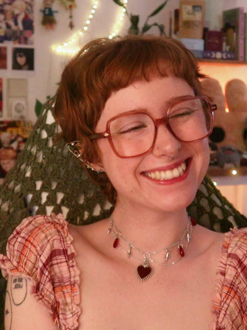 a photo of emmy smiling at the camera. she is wearing silver elf ear cuffs, a silver and red necklace, and big red glasses.