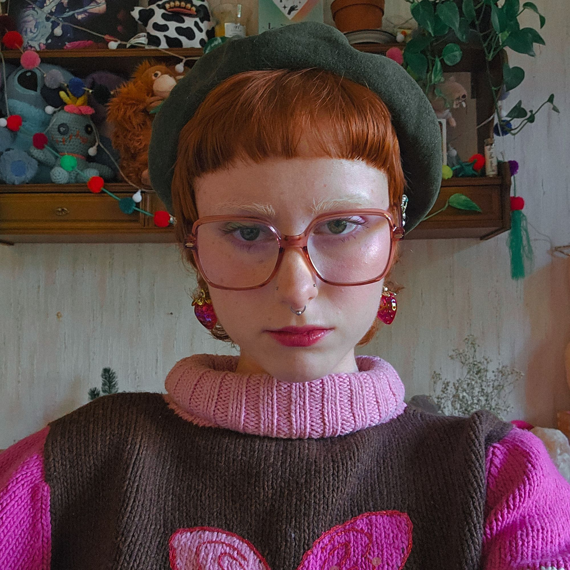 photo of emmy looking at the camera. she is wearing a thick wool, brown and pink jumper with a butterfly on it, a green baret, strawberry earings, and big red glasses.