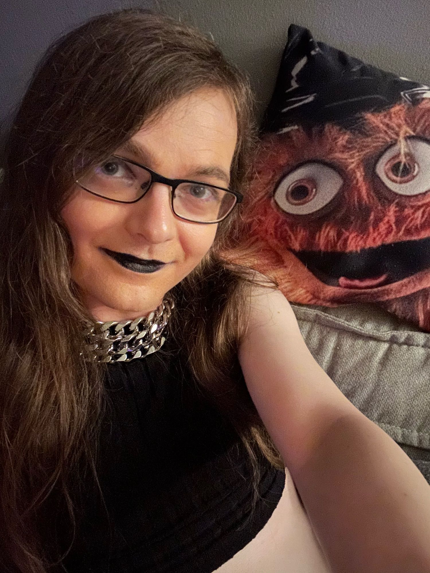 A girl in a black crop top, black lipstick, and black glasses with long brown hair and a chunky layered chain necklace, resting on a couch next to a pillow with the smiling orange furry face of esteemed Philadelphia Flyers mascot, Gritty. She’s grinning a little bit too.