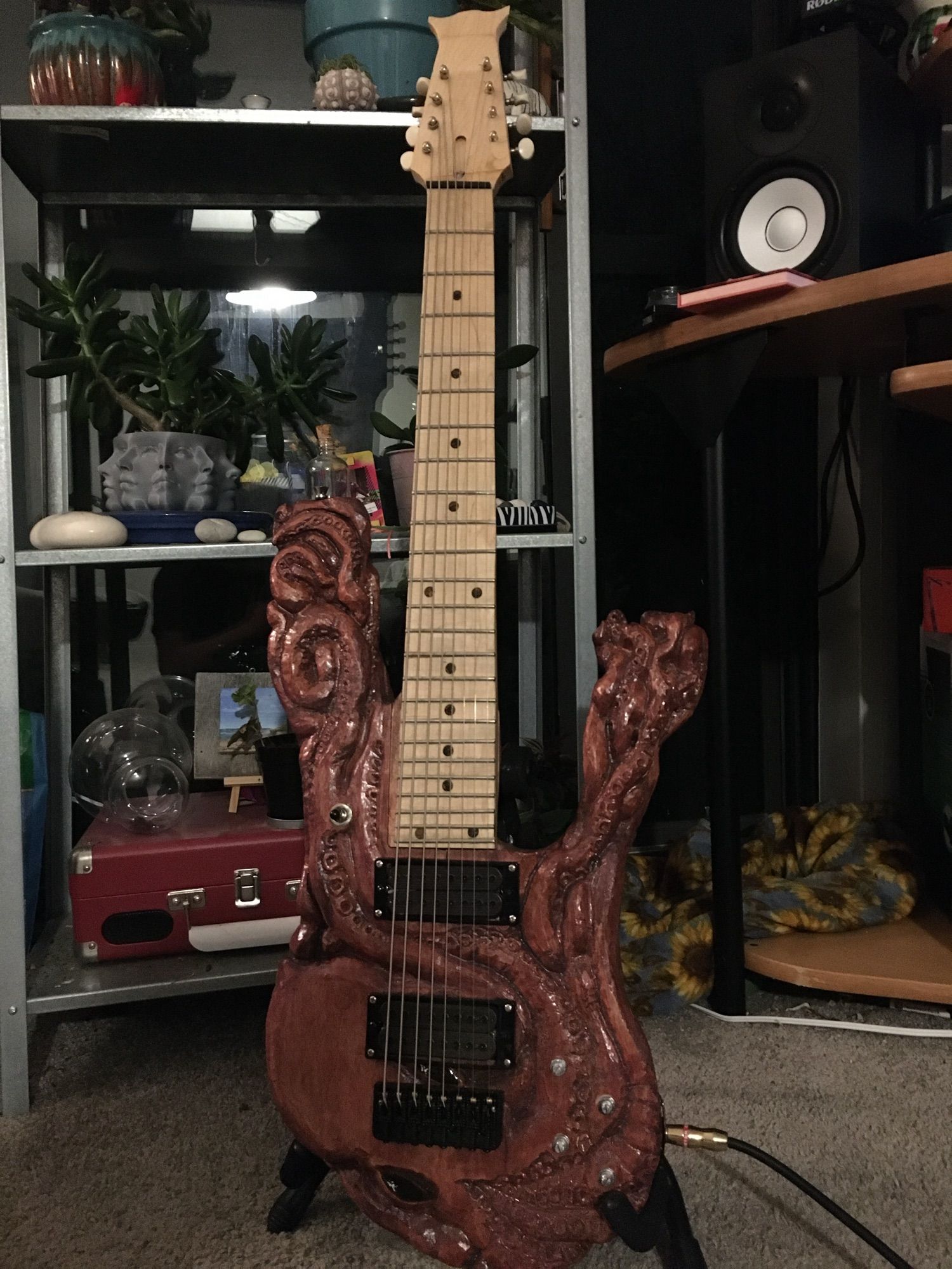 An eight-stringed electric guitar with a reddish-brown body, carved in the shape of a kraken. A Yamaha HS-5 is visible in the background, as well as a metal shelf with an assortment of potted succulents, stones, artwork, and a portable record player.