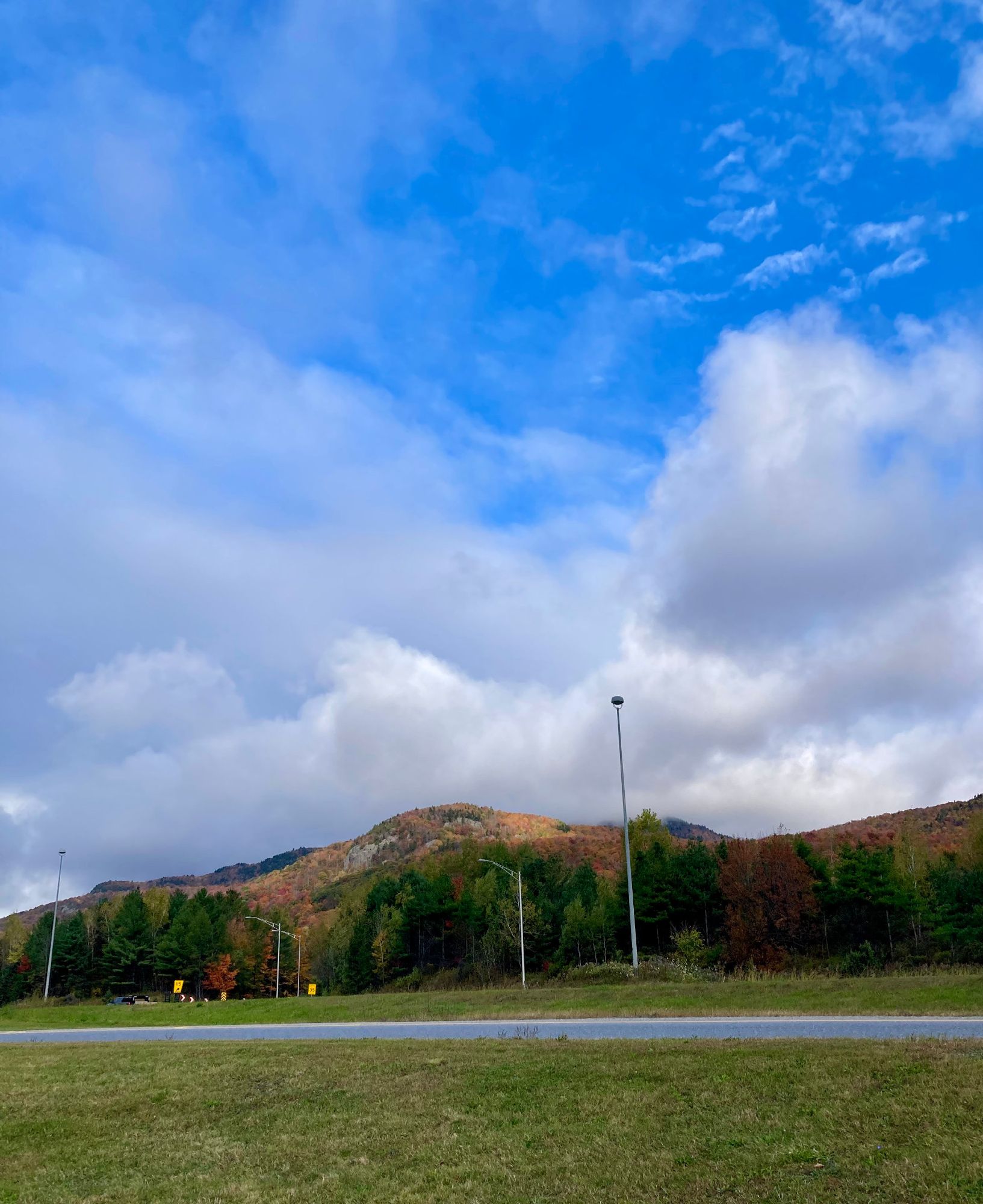 Dans une halte routière, le long d'une autoroute, photo d'une montagne colorée de vert, de jaune, de safran, d'orange et d'un soupçon de rougeurs par-ci, par là sous un ciel bleu pavoisé de nuages qui s'effilochent.