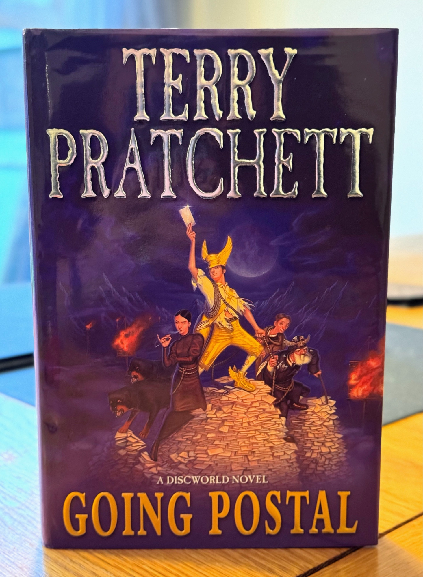 2004 Doubleday copy of Going Postal, first published 25th September, featuring the original ‘Star Wars’ Kidby cover art. Art shows Moist, a tall, dark-haired young man, wearing his tattered golden suit and winged hat, standing on top of a pile of post, holding a letter in the air like a sword. Spike (aka Adora Belle Dearheart) stands below, at his side, smoking. In the background there are burning clacks towers. 

‘It wasn’t a nice thing to do,’ Adora Belle Dearheart went on, in the same level tone. 

‘There wasn’t a nice thing that would work,’ said Moist.