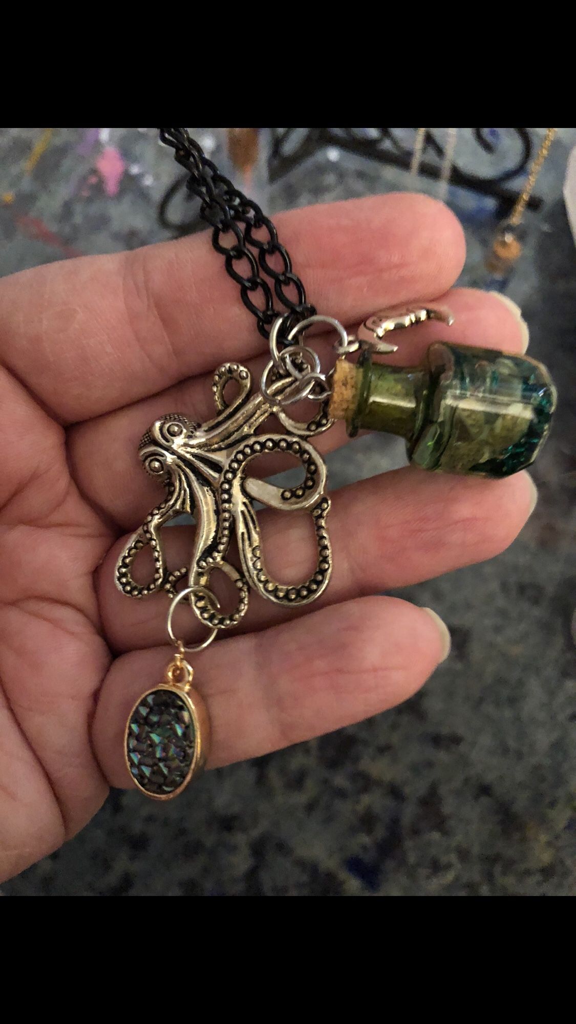 A necklace with two silver moon charms on either side of a silver octopus pendant. Attached to one of the octopus’s arm is a greenish geode like charm and towards the upper part is a transparent green bottle with green beads and brown seashells