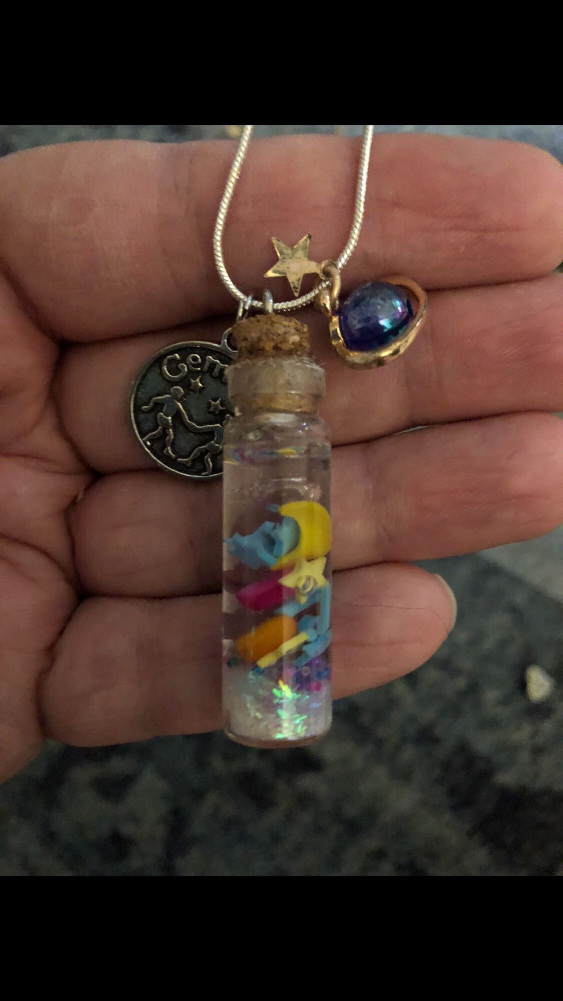 A necklace with a silver medallion that says Gemini next to a bottle filled with glitter and small hearts, stars and a crescent moon and next to that is a small iridescent planet surrounded by a golden ring charm