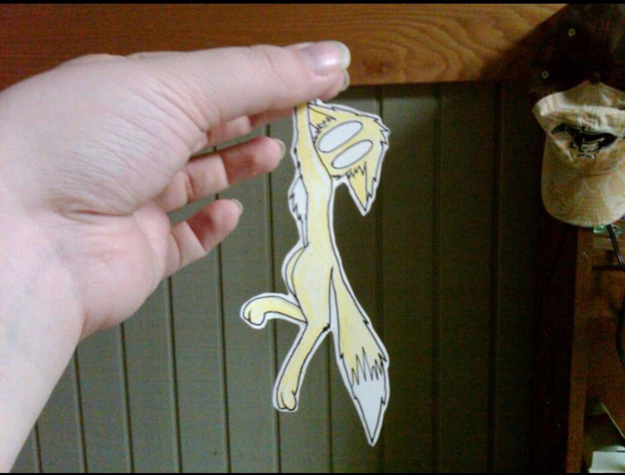 A photo of a person’s hand holding a cut out drawing of a yellow cat as if the cat is dangling from the person’s grip. A concerned look is on the cats faces as she looks down