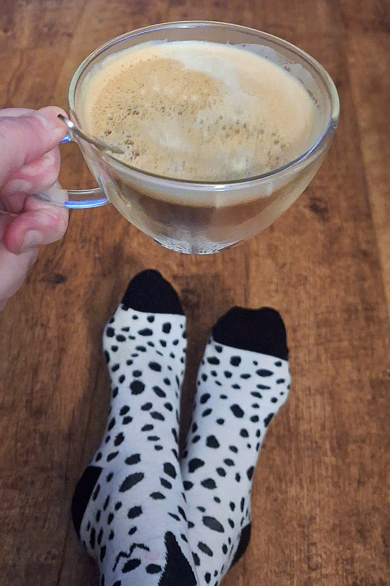 M'n hand houdt een dubbelwandig glas vast met koffie en een lepeltje, daaronder m'n voeten met sokken: wit met kleine zwarte vlekken (partoon van een dalmatiër), zwarte hiel en teen. Boven de enkel is een hondenkop te zien. (Dalmatiër)