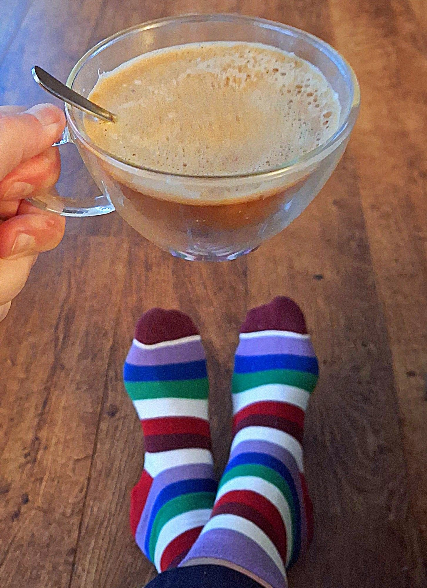 Een hand die een dubbelwandig glas met koffie bij het oor vasthoud, met op de achtergrond voeten die donker rood, wit, lila, blauw, groen, rood gestreepte sokken dragen.