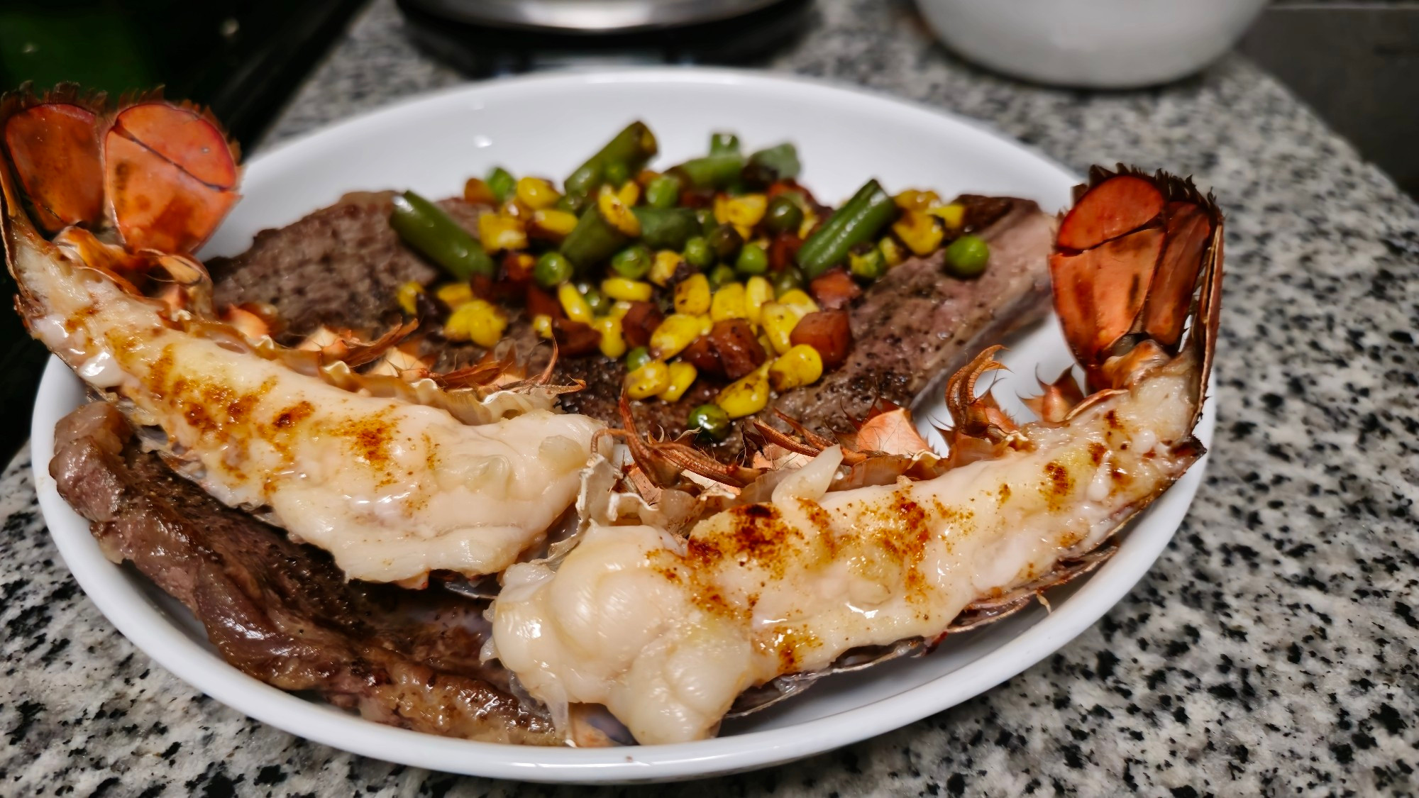 A surf-and-turf plate, with a 3 oz lobster tail split in 2 length-wise, bone-in steak, and mixed vegetables.