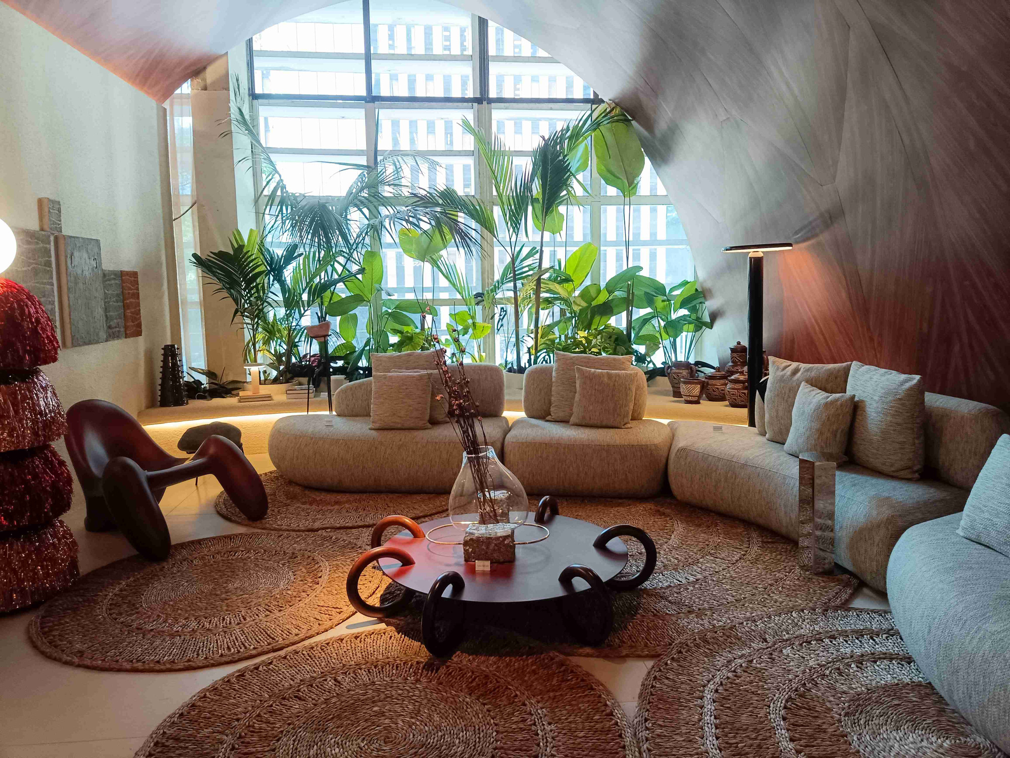 Portuguese: Imagem de uma sala onde o teto em formato circular e a mesa com pés em formato de anel chamam a atenção. Ainda é possível ver sofás confortáveis e uma ampla janela com plantas naturais.

English: Image of a living room where the circular ceiling and the table with ring-shaped frames draw attention. It also features comfy sofas and a large window with natural plants.