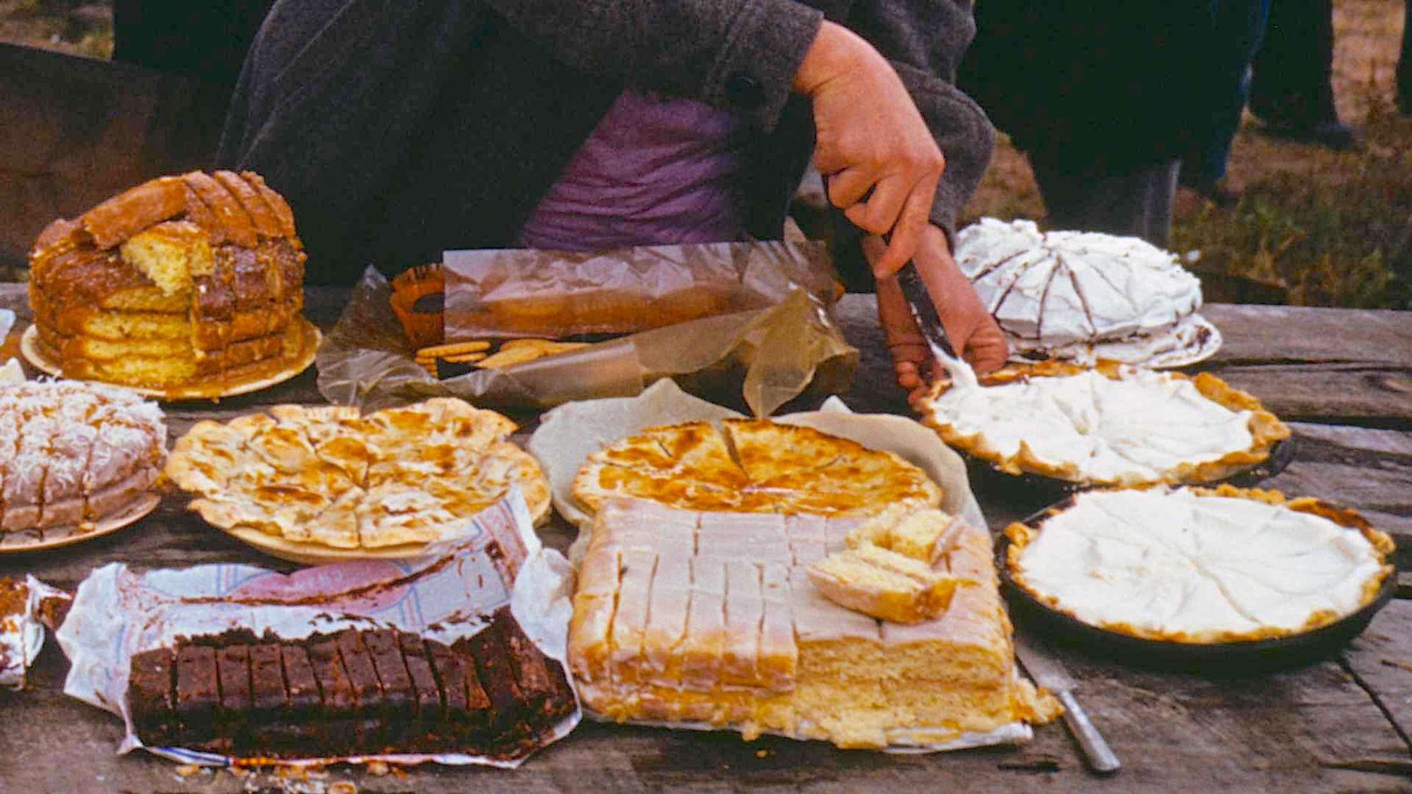 A variety of homemade baked goods on a table, including round pies with meringue toppings, a layered cake, and chocolate bars. A person is cutting a pie. Arrangement suggests an outdoor event or gathering focused on sharing food.