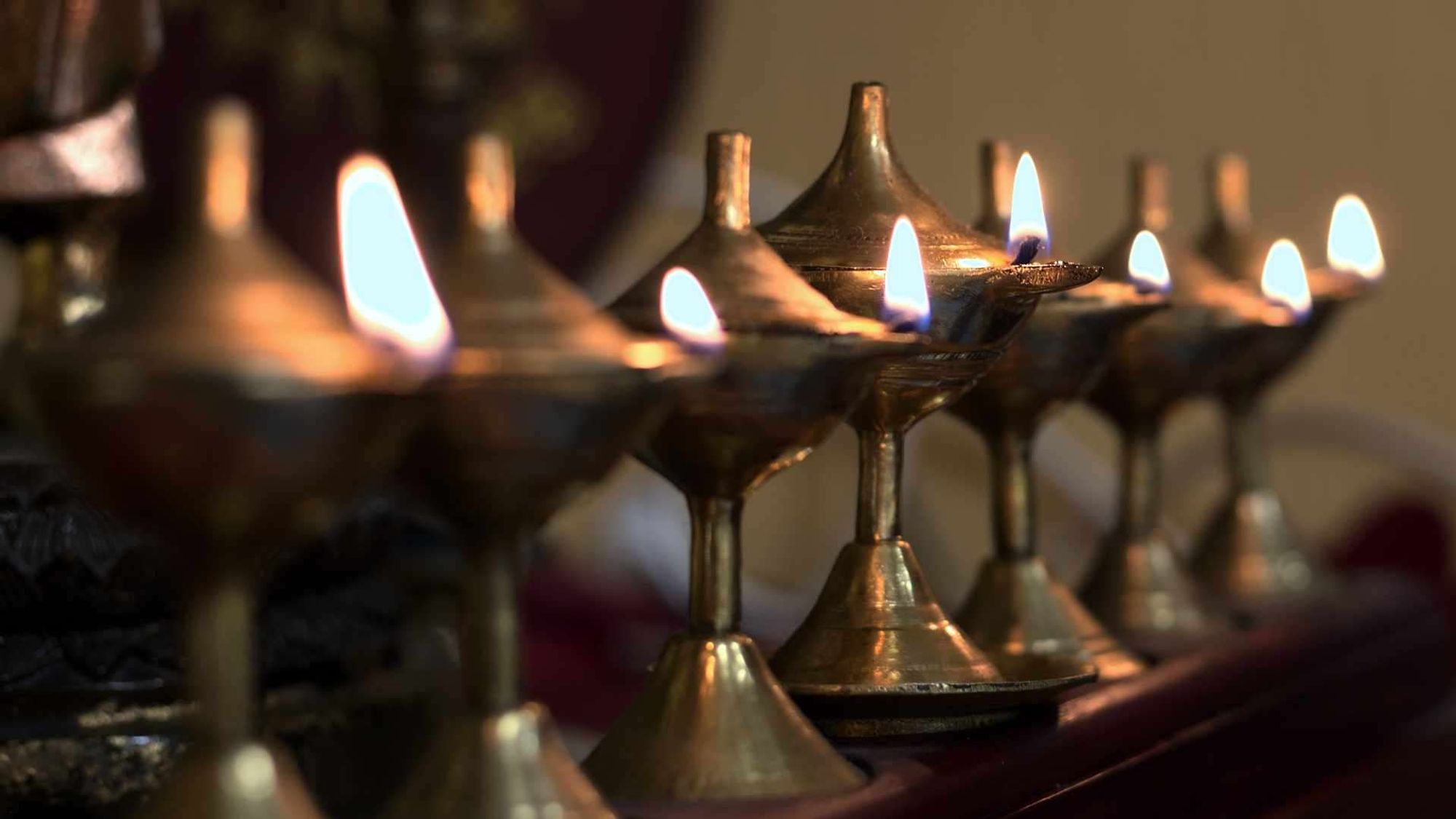 A row of lit oil lamps with glowing flames. The focus is on the nearest lamps, with a blurred background enhancing the warm glow.