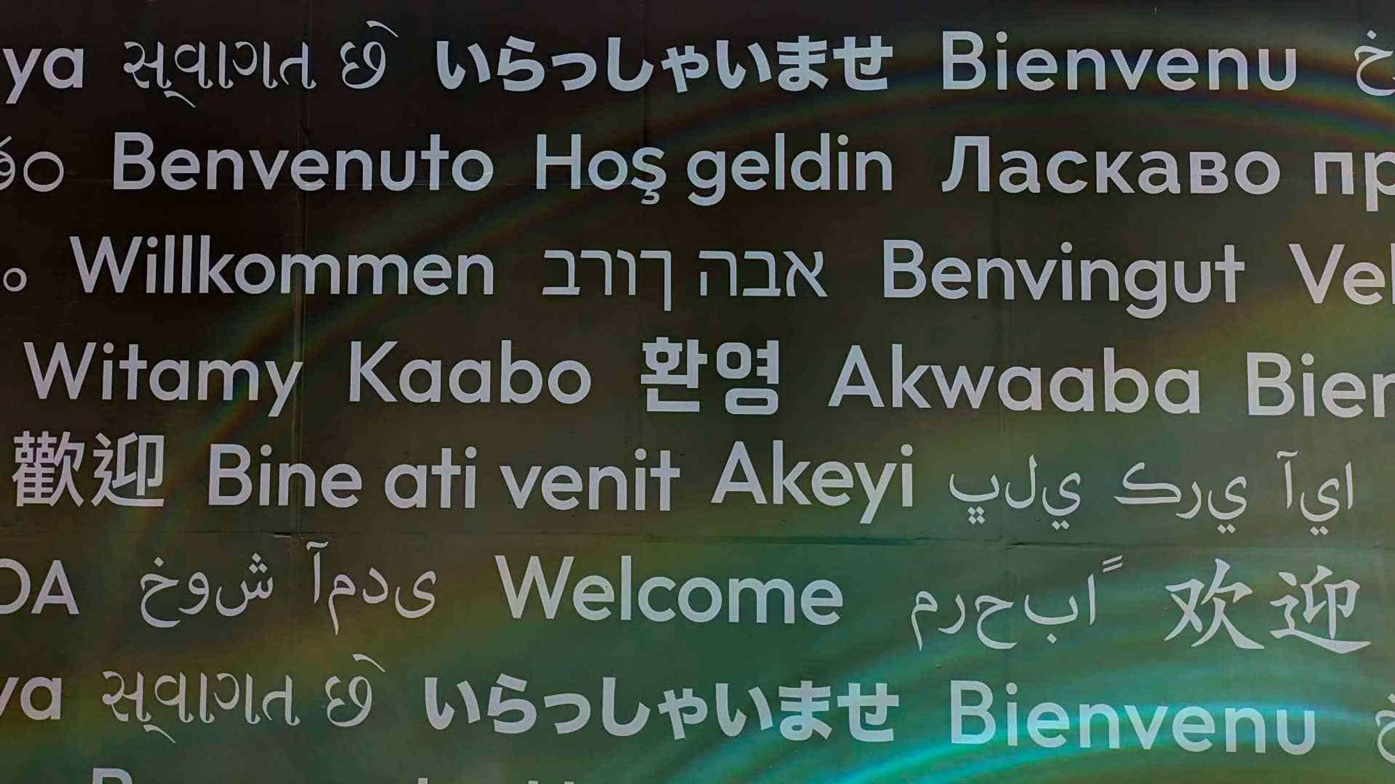 The image displays the word “Welcome” in various languages and scripts, arranged in a colorful, visually appealing manner against a dark background.