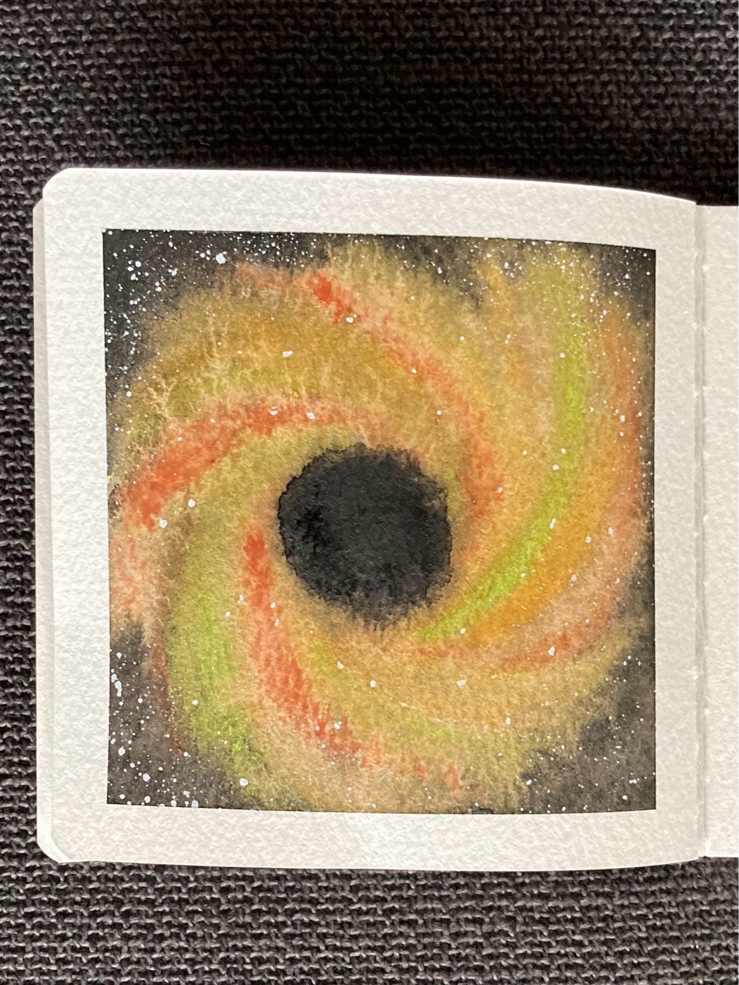 A watercolor painting of a black hole in space. The center of the black hole is black and completely devoid of stars. Around the center is a swirling halo of yellow with green and orange-red stripes. Both the halo and the surrounding space are dotted with stars.