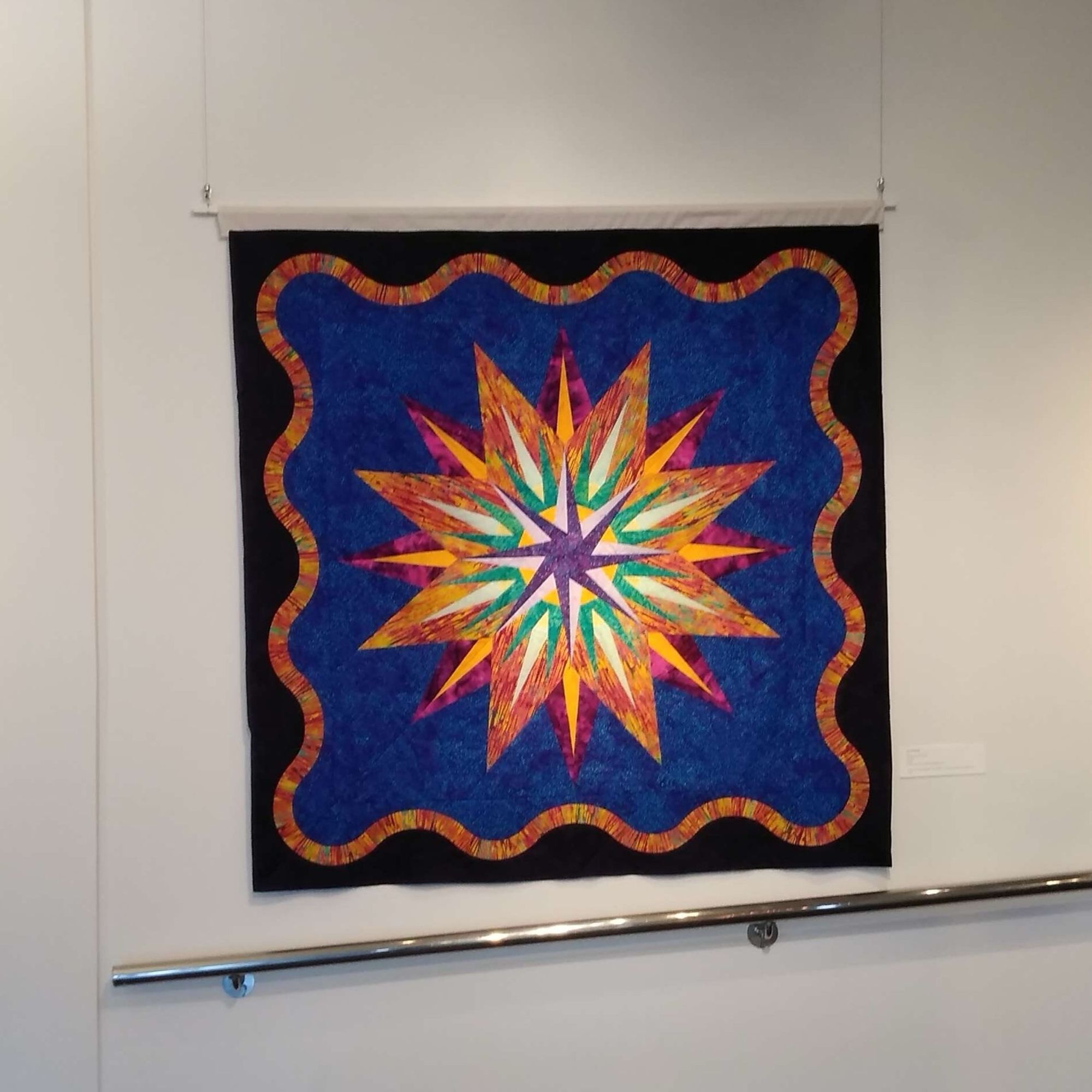 A 62“ x 62” square quilt hangs on the white wall of a gallery over a handrail. It has a bright central mariner’s compass made of orange, turquoise, purple, and yellow batiks on a background of vivid cobalt blue, surrounded by a curvy orange batik border, with a frame of dark purple eggplant.