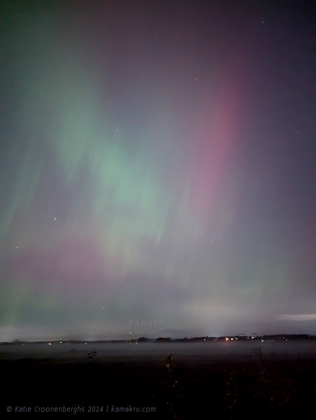 A photo of the northern lights in soft colors with fog in the field and some lights in the distance. Text reads copyright Katie Croonenberghs 2024 kamakru.com