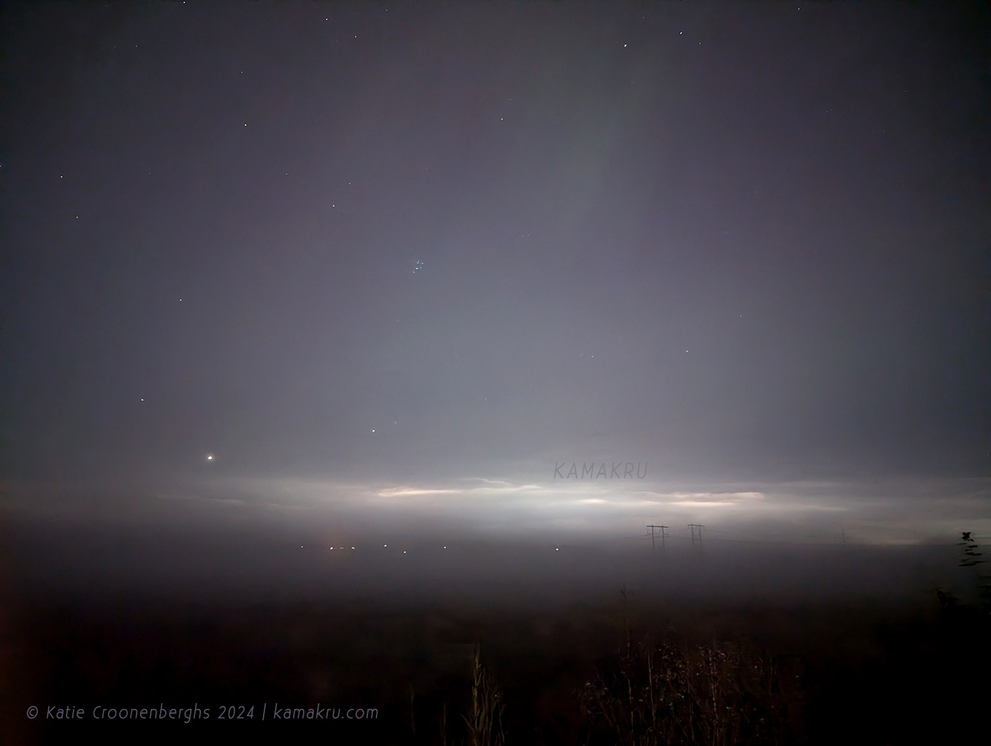 A photo of the night sky with a hint of a green Aurora through the fog and clouds with bushes in the foreground. Text reads copyright Katie Croonenberghs 2024 kamakru.com