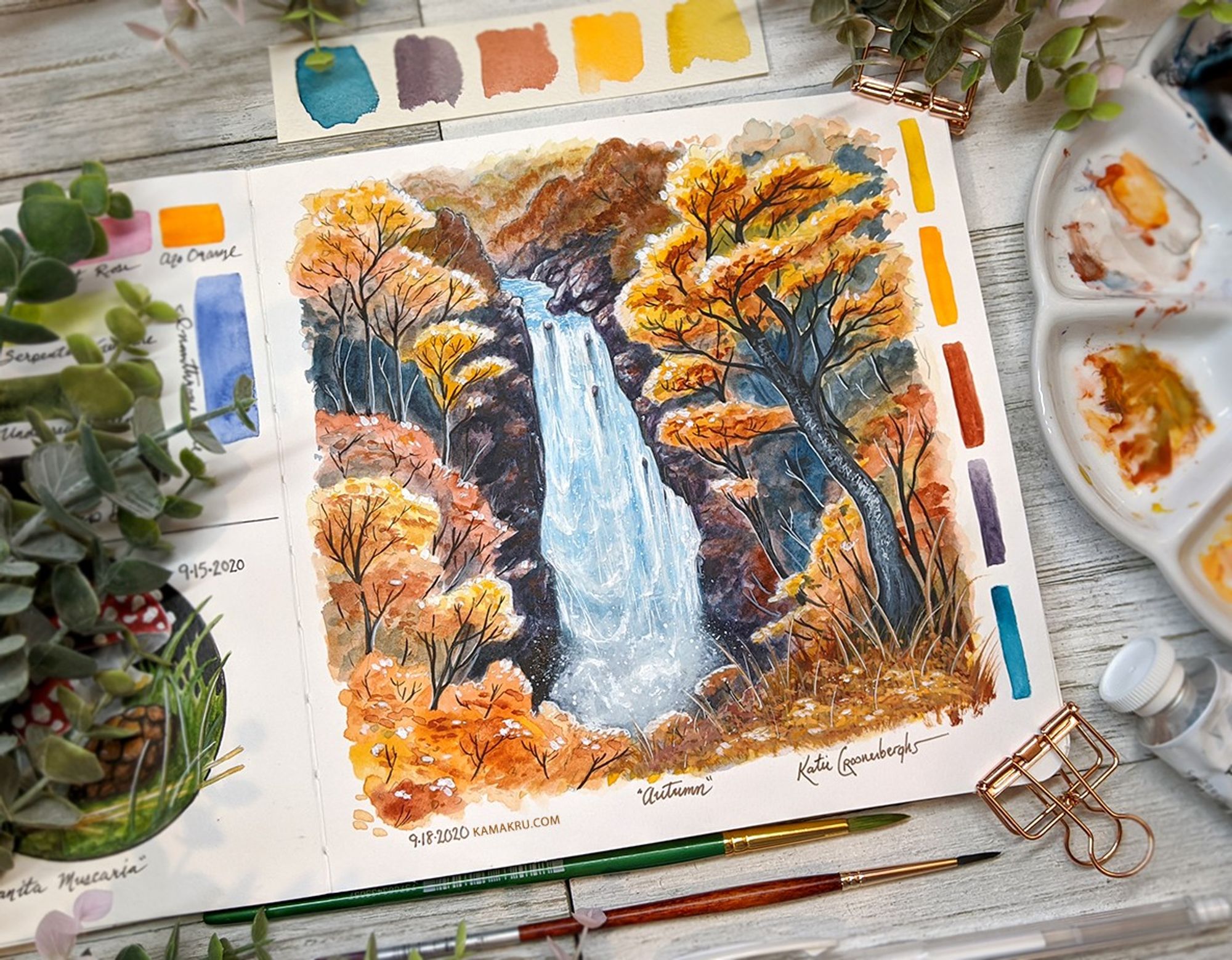 Painting in a sketchbook of a blue waterfall framed by bright orange autumn trees. Watercolor and gouache. The sketch is in a book that sits flat on a table. At the right is part of the artist's paint palette, to the left are some decorative leaves. Written under the painting is the date, 9-18-2020 kamakru.com the title "Autumn" and signature.