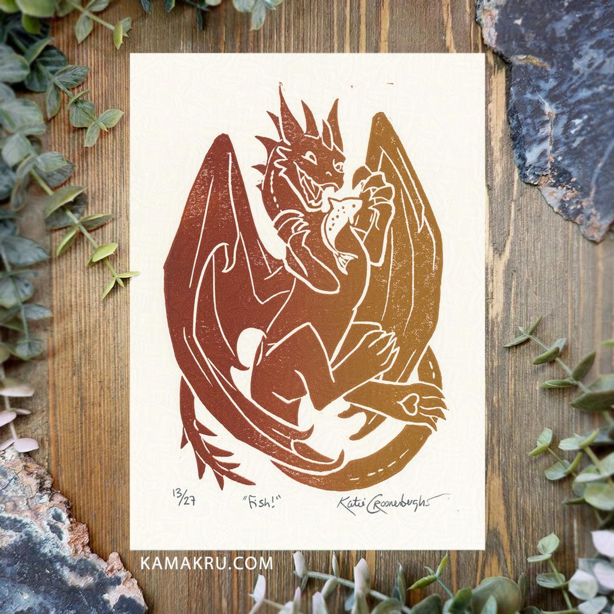 A two colored print of a reddish clay color and muted yellow. A cute dragon plays with a fish. It's a limited edition print. The edition number of this print is 13/27, the title is written in the center "Fish!" and signed Katie Croonenberghs. Text under the print reads kamakru.com