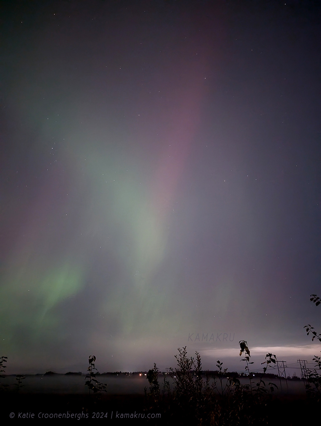 A photo of the northern lights in soft colors with bushes in the foreground, fog and some lights in the distance. Text reads copyright Katie Croonenberghs 2024 kamakru.com