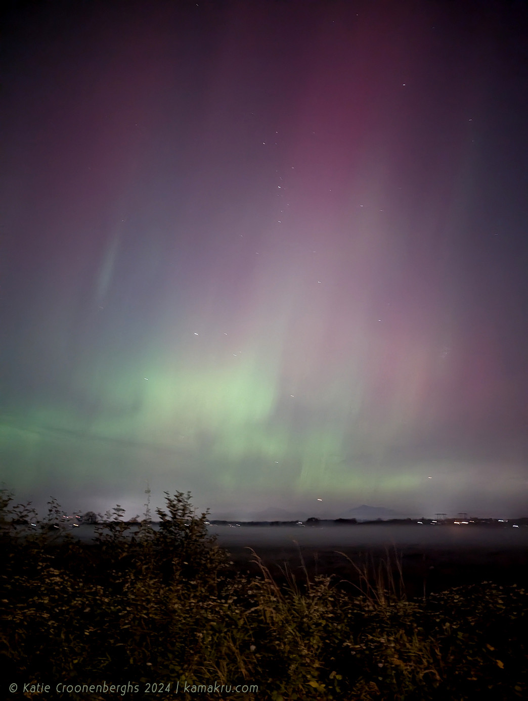 A photo of the northern lights in soft colors with bushes in the foreground, fog and some lights in the distance. Text reads copyright Katie Croonenberghs 2024 kamakru.com