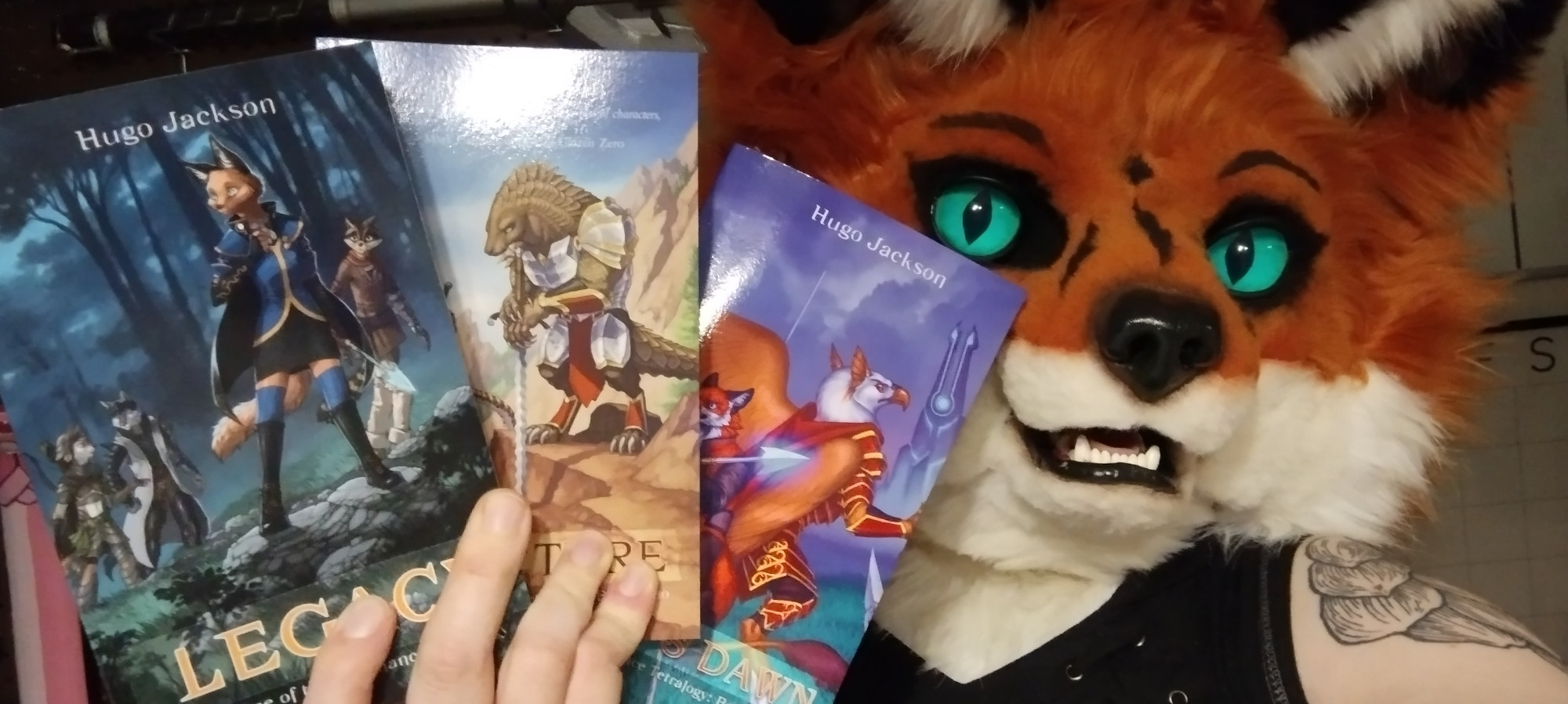 A fox fursuiter holding a set of three young adult first fantasy novels: the
first trio of books in the Resonance Tetralogy series