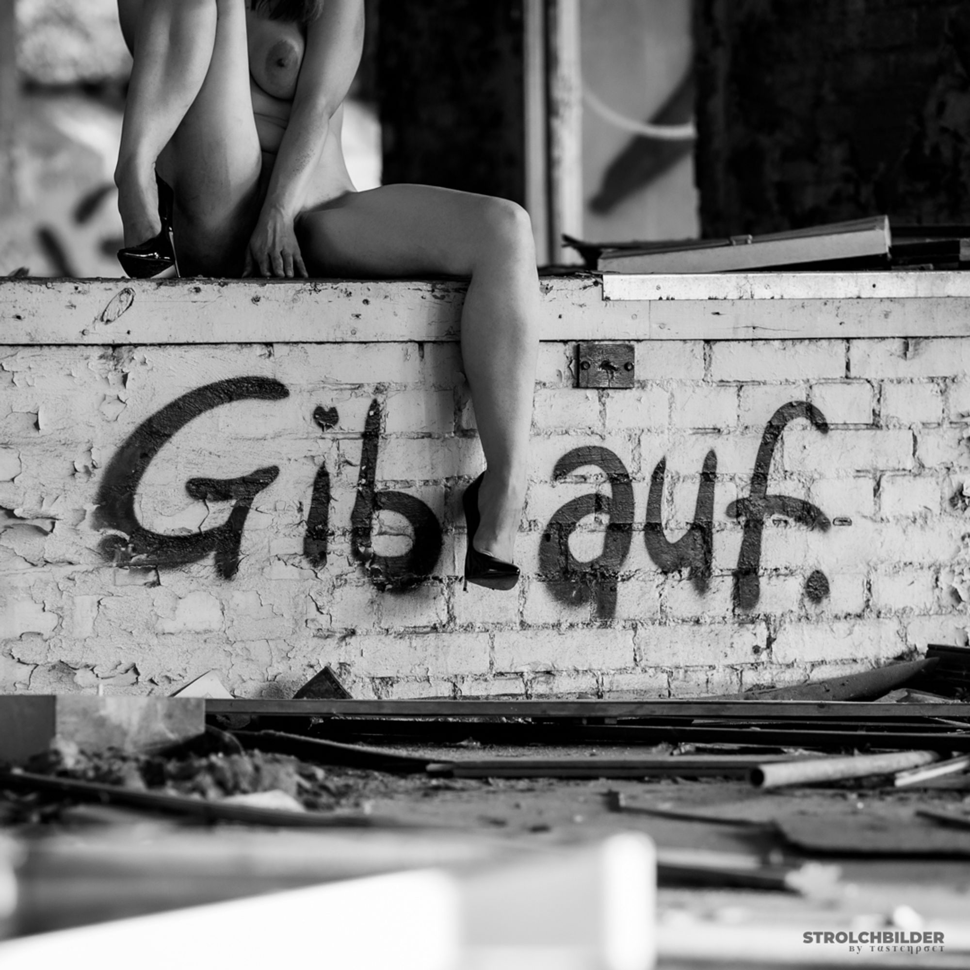 Teilakt, die Person sitzt auf einer Mauer und trägt Stilettos mit Metallabsatz. Auf der Mauer aus Backsteinen steht als Grafitto "Gib auf." Ein Bein hängt zwischen den Wörtern von der Wand.