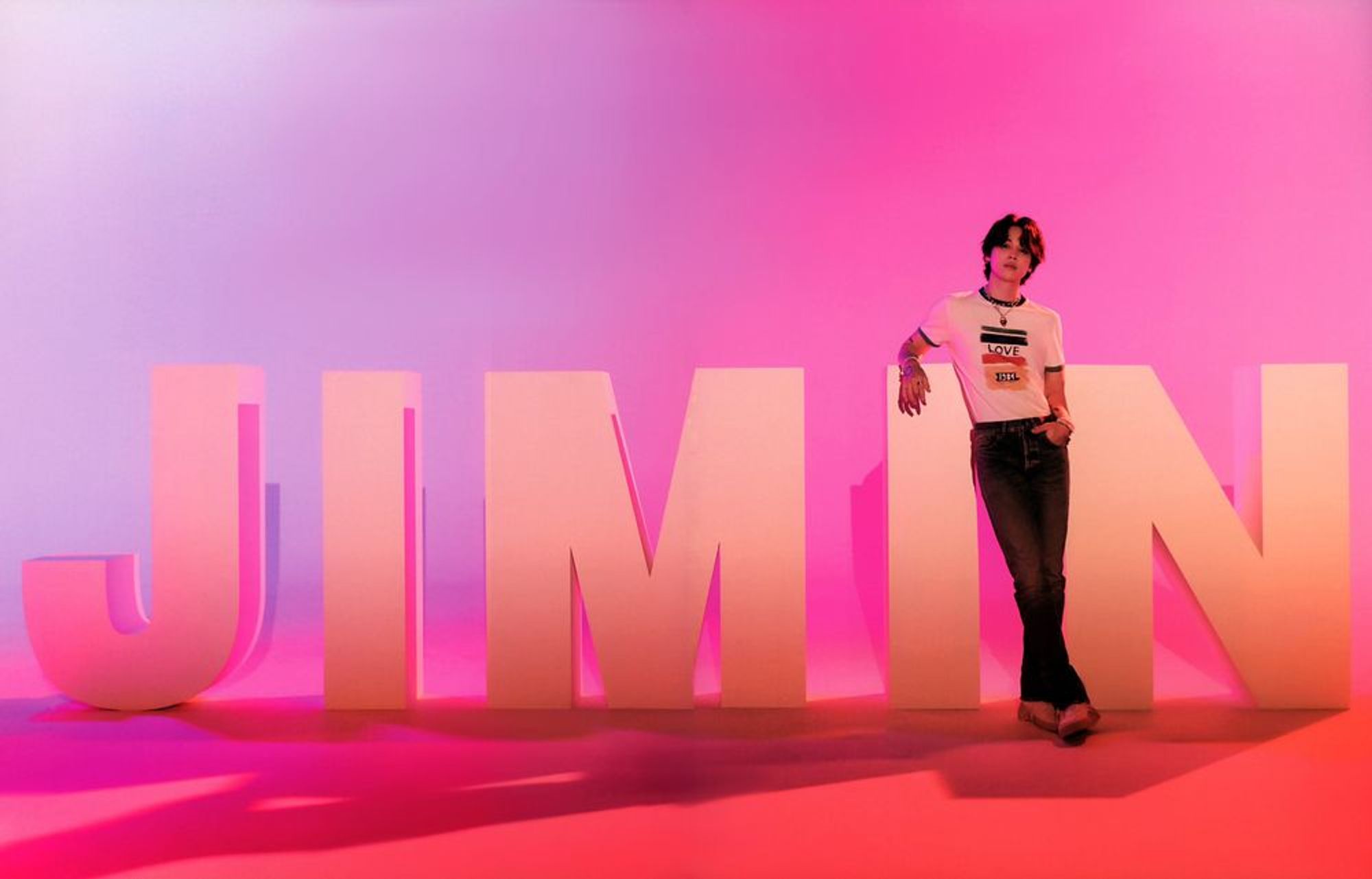 Jimin in a tshirt in jeans leaning against a large letter I. His name is spelled out behind him in large white laters against a pinkish-orangey background