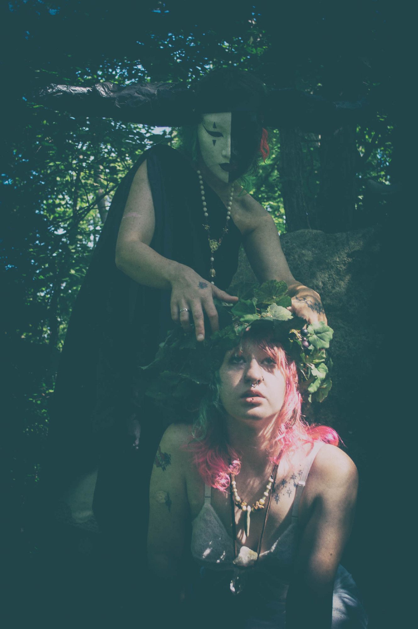 matte color photo. a shadowy horned and masked figure in a black chiton looms behind a kneeling young woman with pink hair and a crown of grape vines. The figure is reaching down to the woman's head even as the woman's eyes turn heavenward.