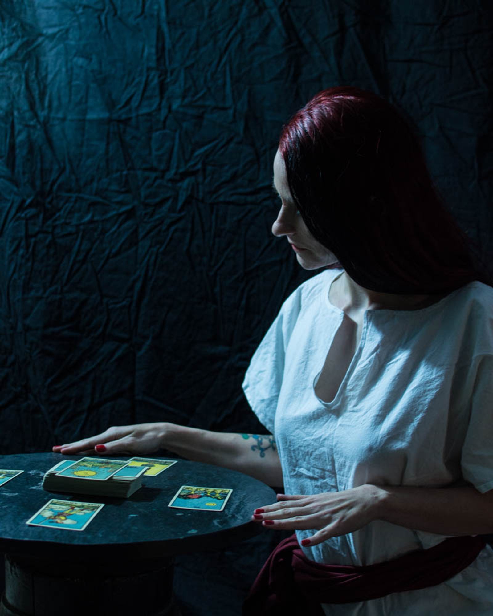 a scrawny white woman with long dark hair sits at a table covered in tarot cards. she is wearing a white robe and a red sash