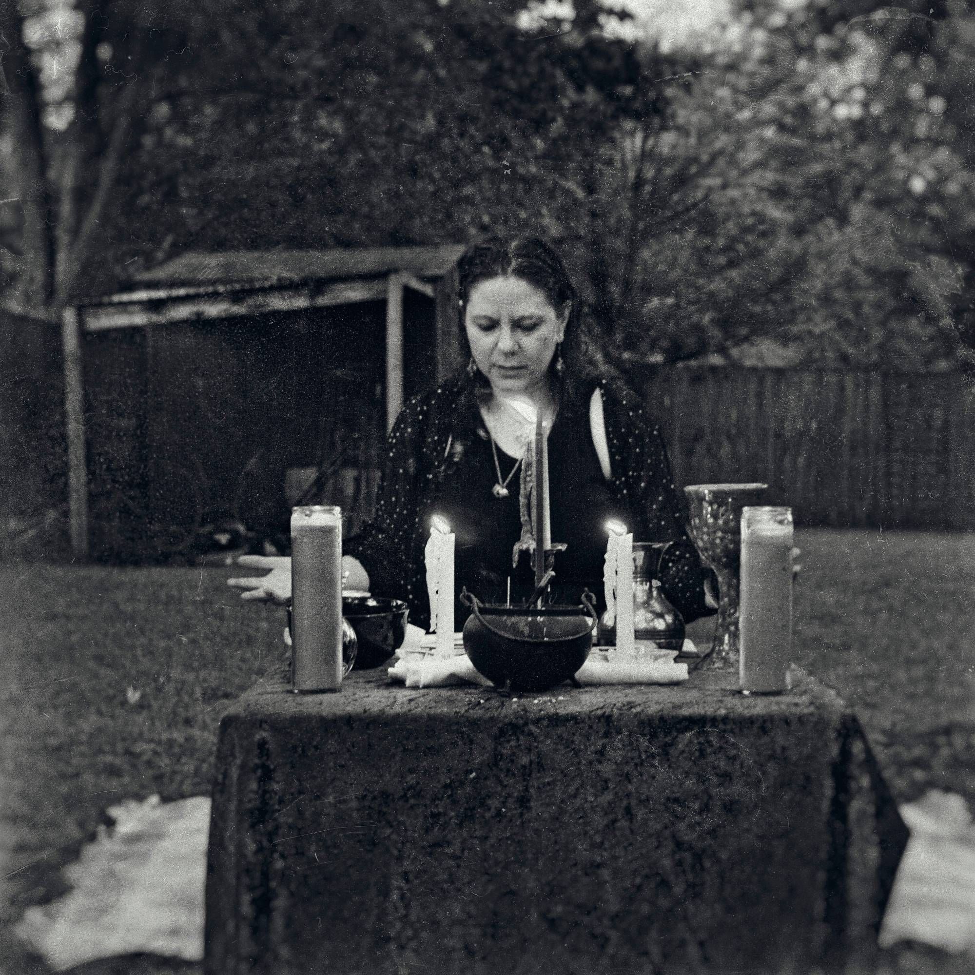 black and white photo of a white woman with dark hair and clothes sitting behind a candlelit altar. a splash of light appears off-center of the frame, a classic orb indicating supernatural activity