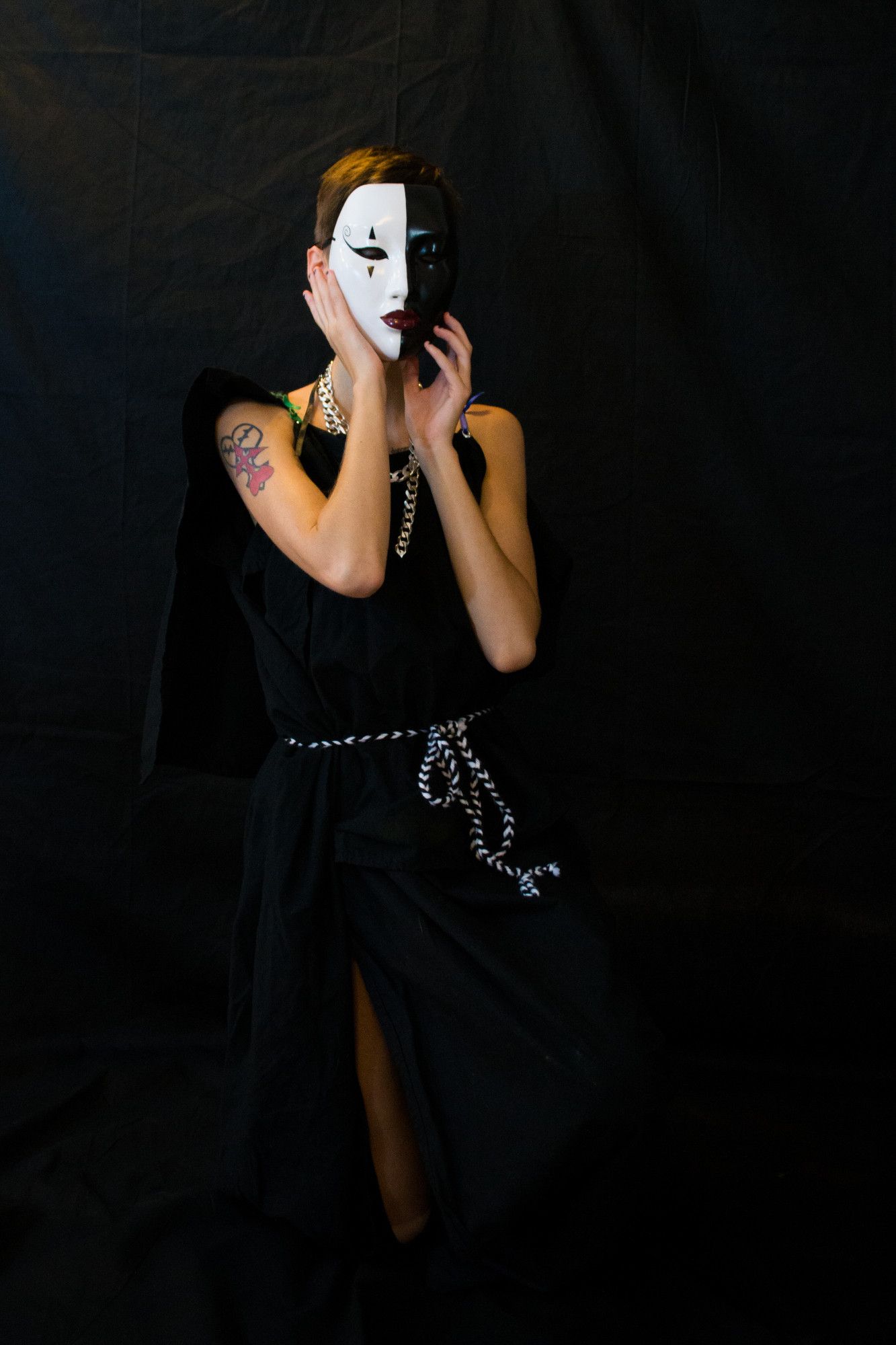 a skinny young white woman with short dark hair dressed in a black chiton bound by a black and white belt and wearing heaving silver chains. she is donning a black and white mask with dark red lips